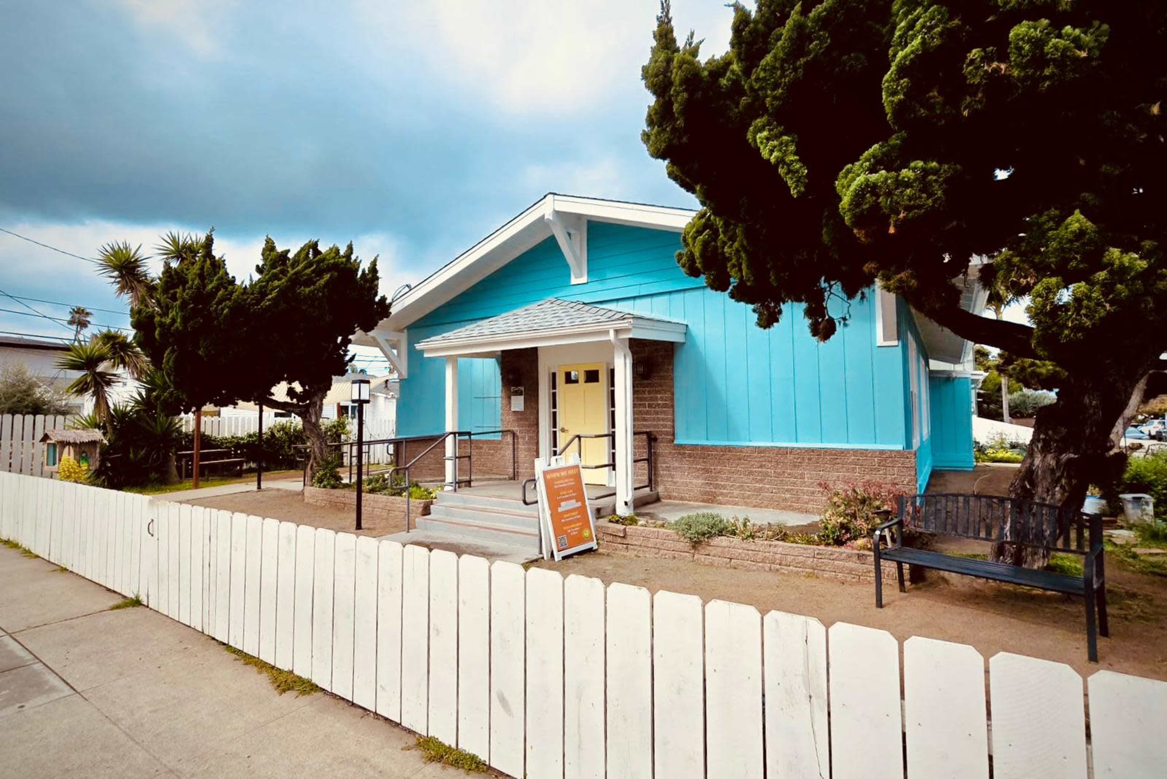 A light blue house with a porch and a white picket fence, surrounded by trees and landscaping.