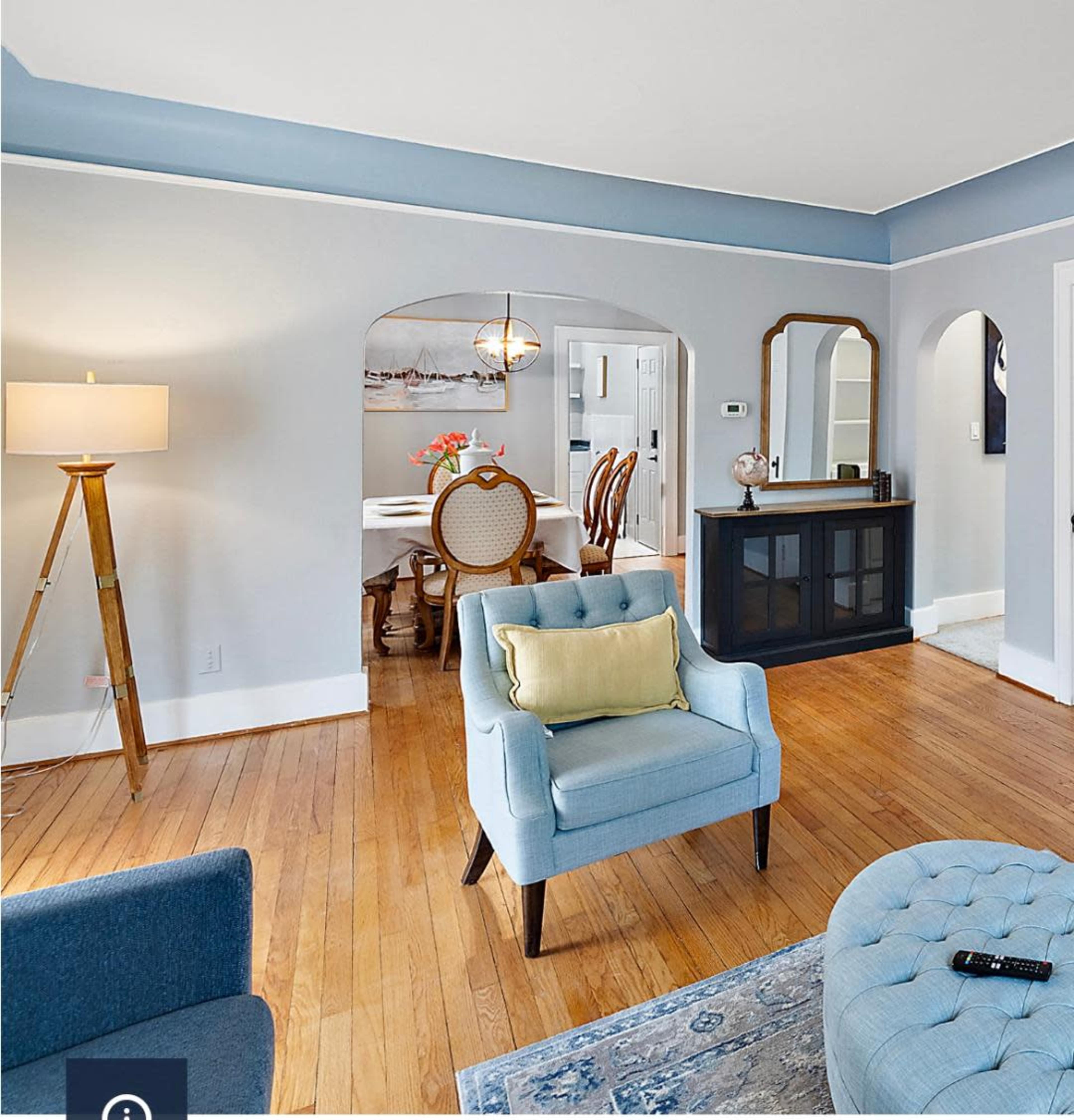 A living room with light blue walls, hardwood flooring, a blue chair with a cushion, and a dining area visible through an archway.