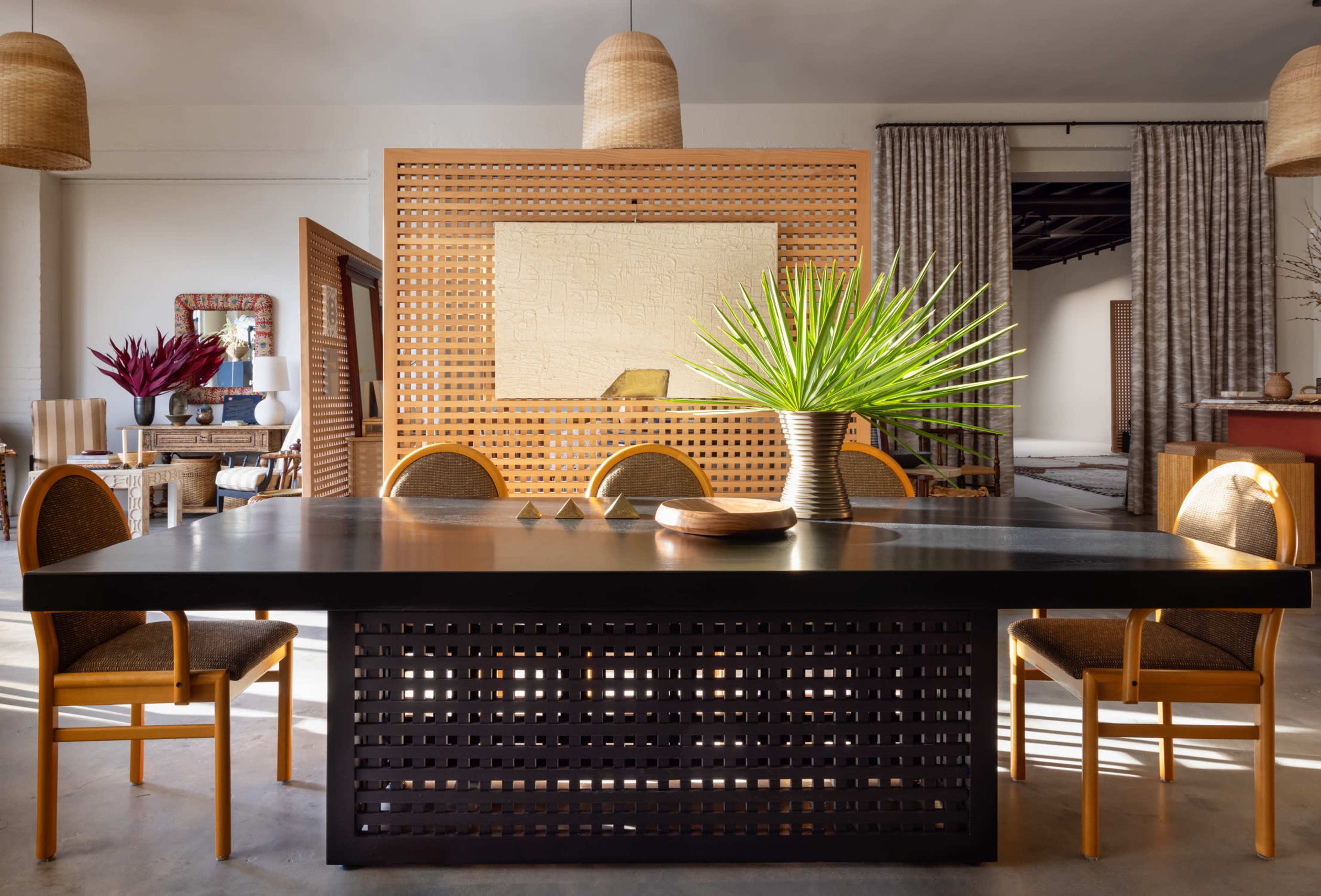 A large black table surrounded by six chairs is set in a well-lit dining area, featuring a decorative screen and a potted plant as the centerpiece.
