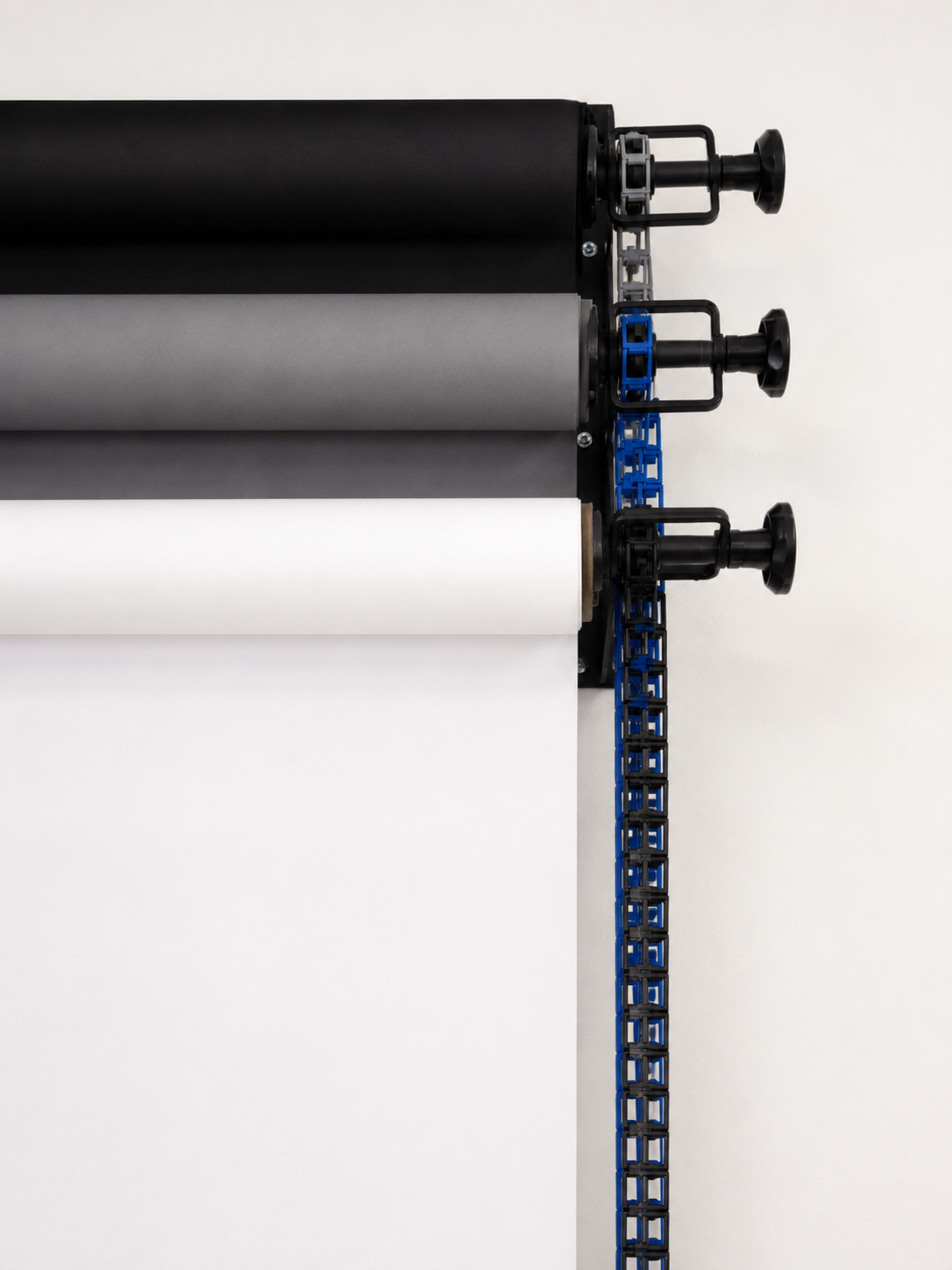 Three rolls of paper in black, gray, and white are mounted on a wall rack with adjustable knobs for rolling.