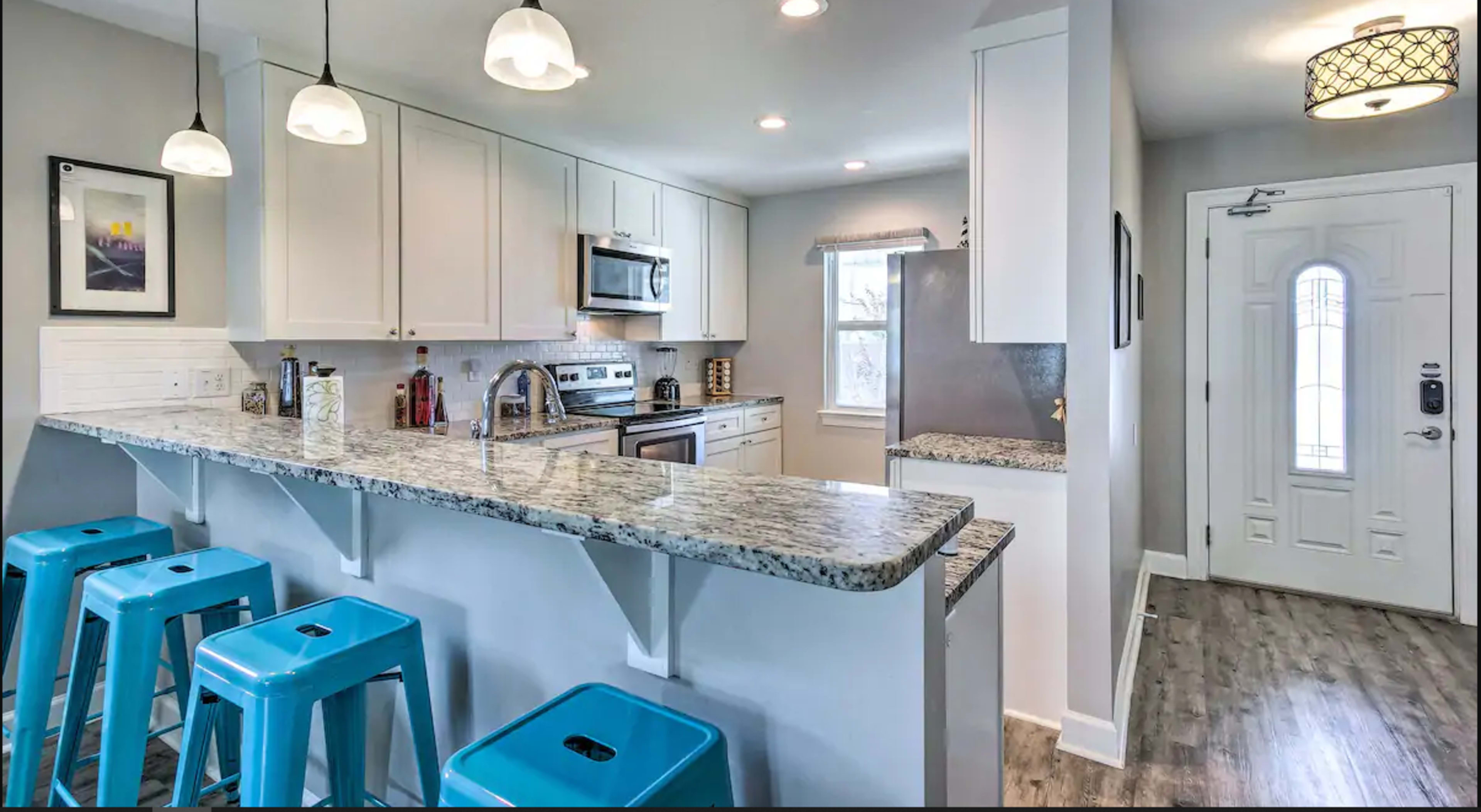 A modern kitchen with white cabinetry, a granite countertop, stainless steel appliances, and blue metal bar stools at a breakfast bar.