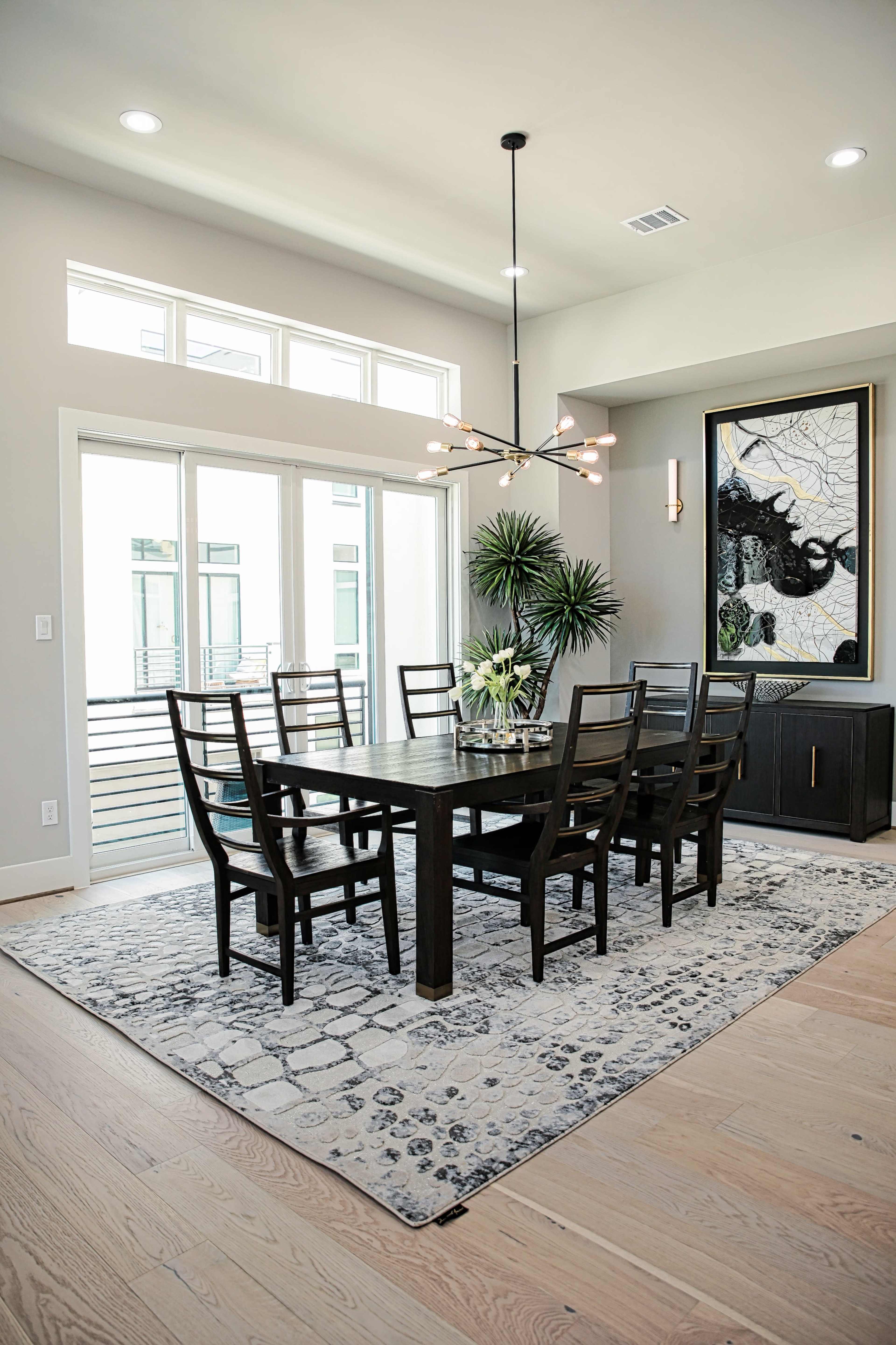 A modern dining room features a large wooden table surrounded by eight chairs, with a decorative rug and a large piece of wall art.