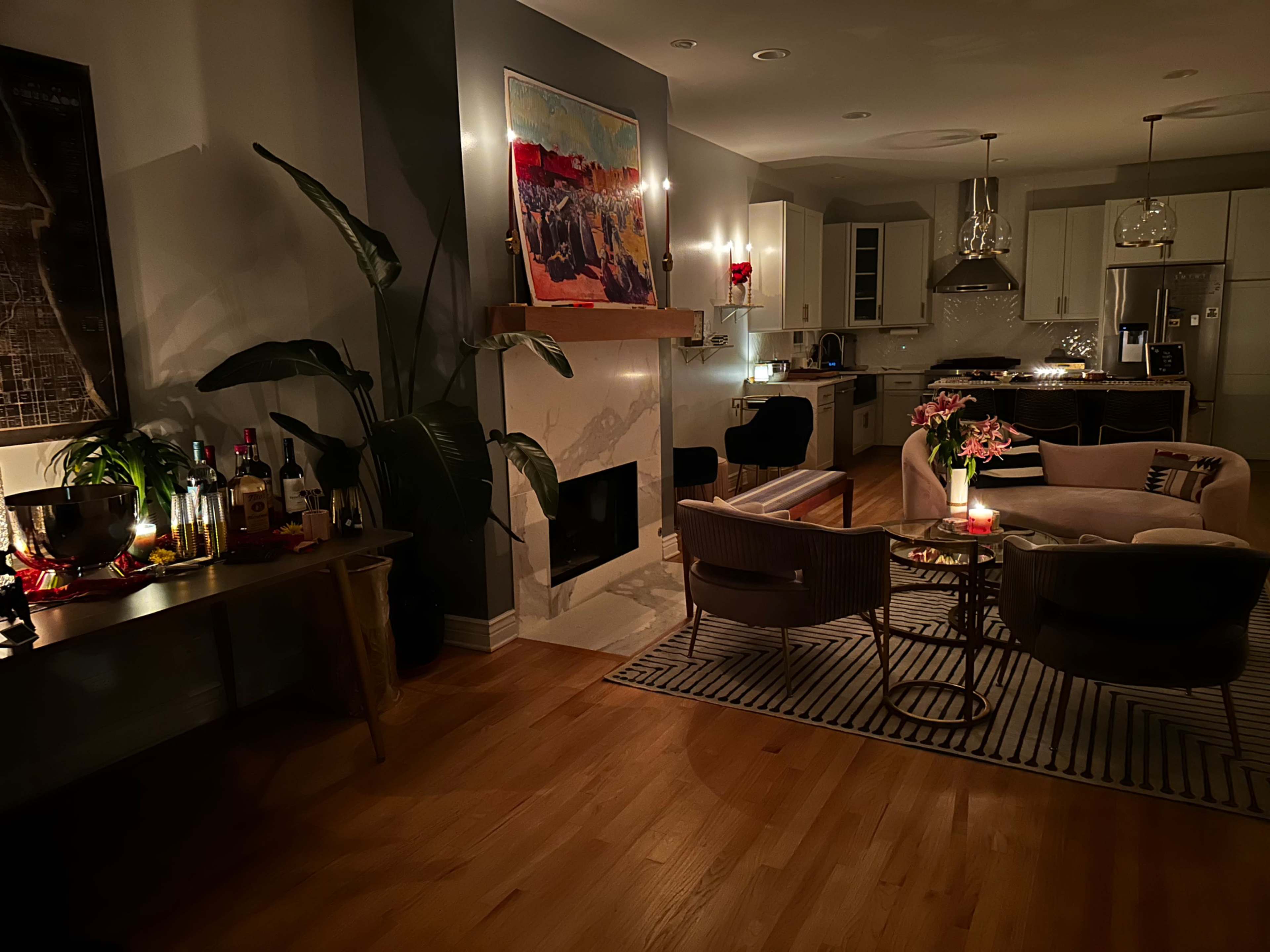 The image shows a cozy living room with a fireplace, comfortable seating, and a table adorned with drinks and flowers, illuminated by soft lighting.