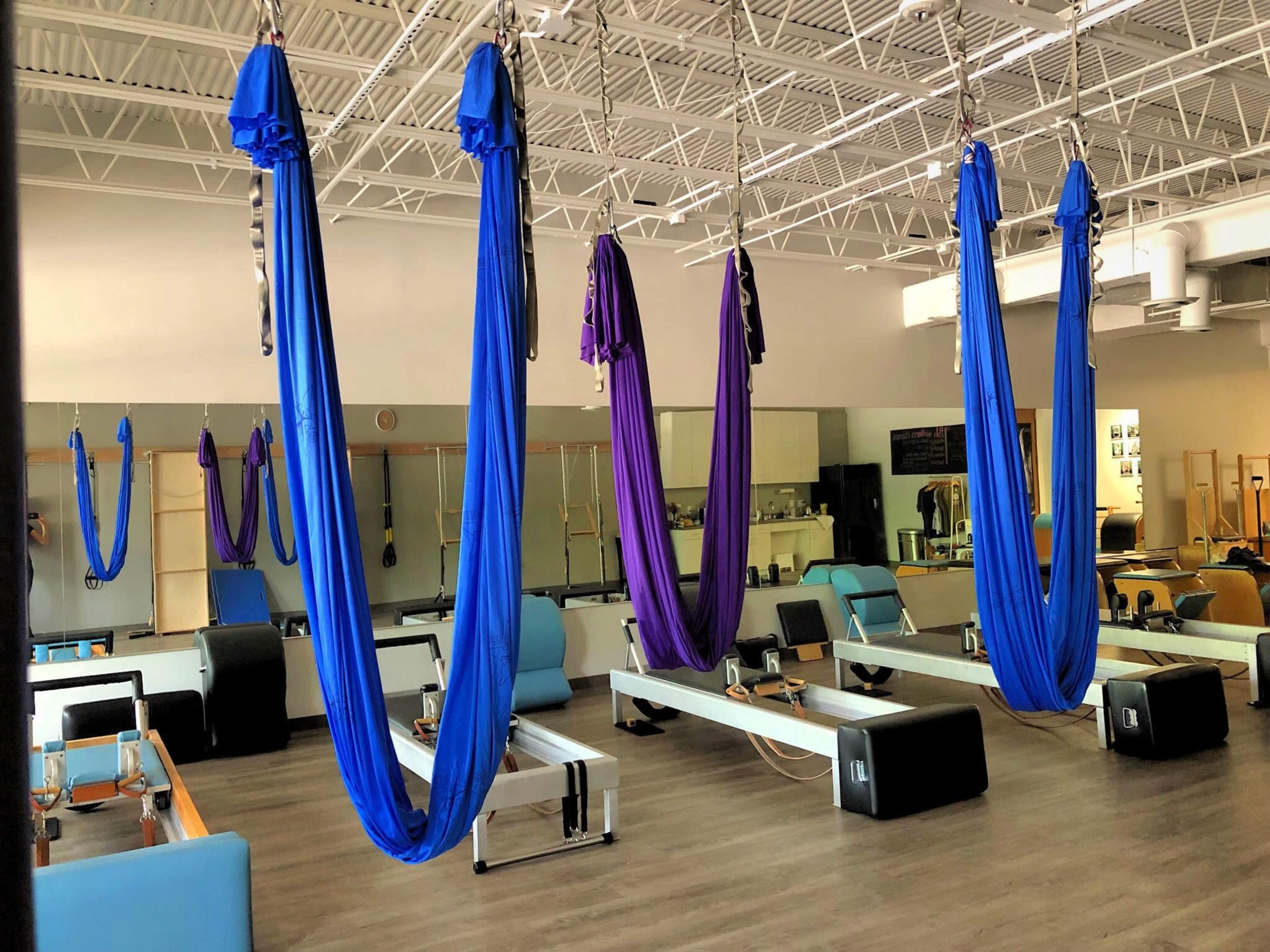 The image shows a fitness studio equipped with aerial hammocks in blue and purple hanging from the ceiling, with exercise platforms arranged beneath them.