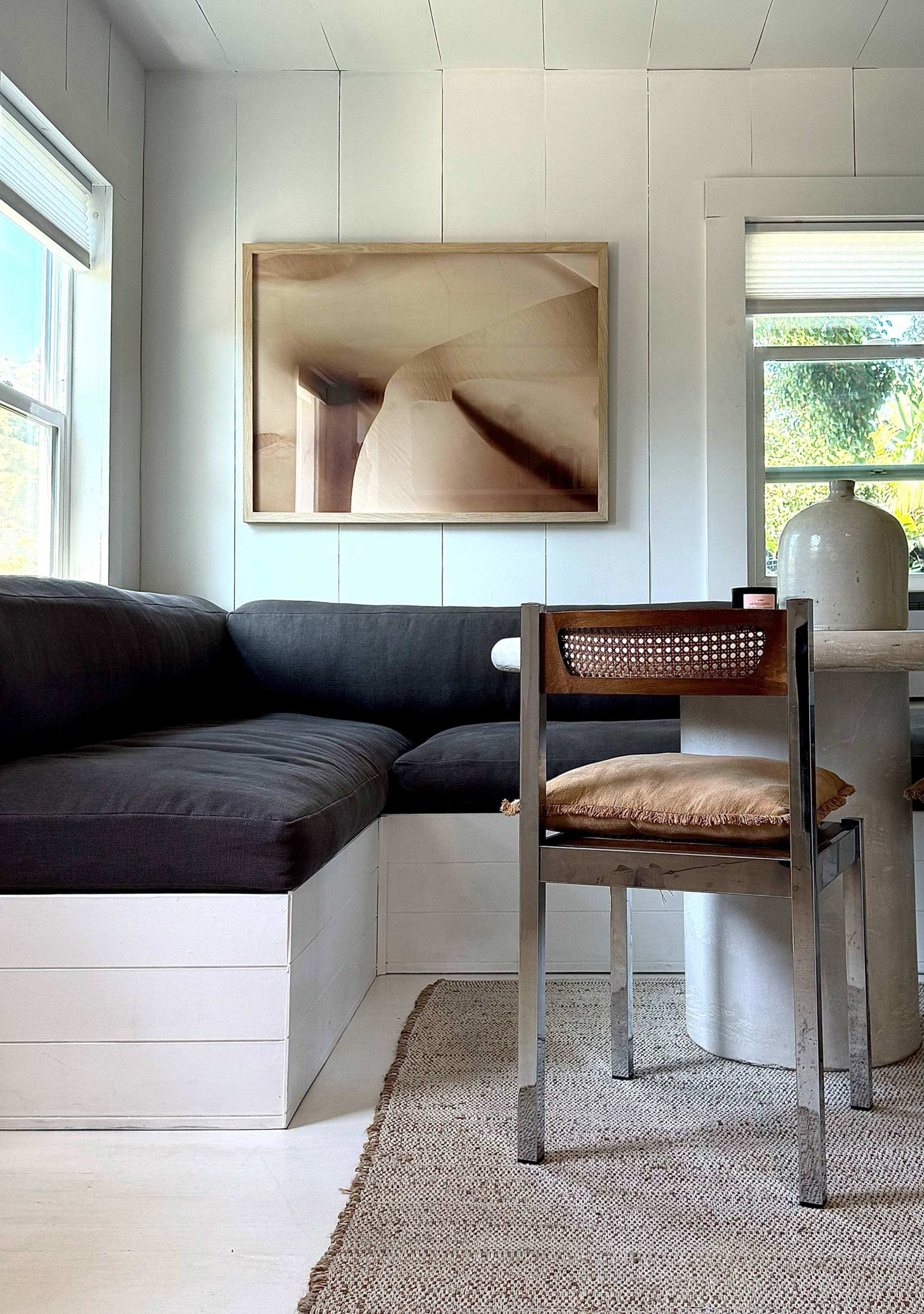 The image depicts a cozy interior with a dark couch, a wooden chair, a round table, and an artwork hanging on the wall.