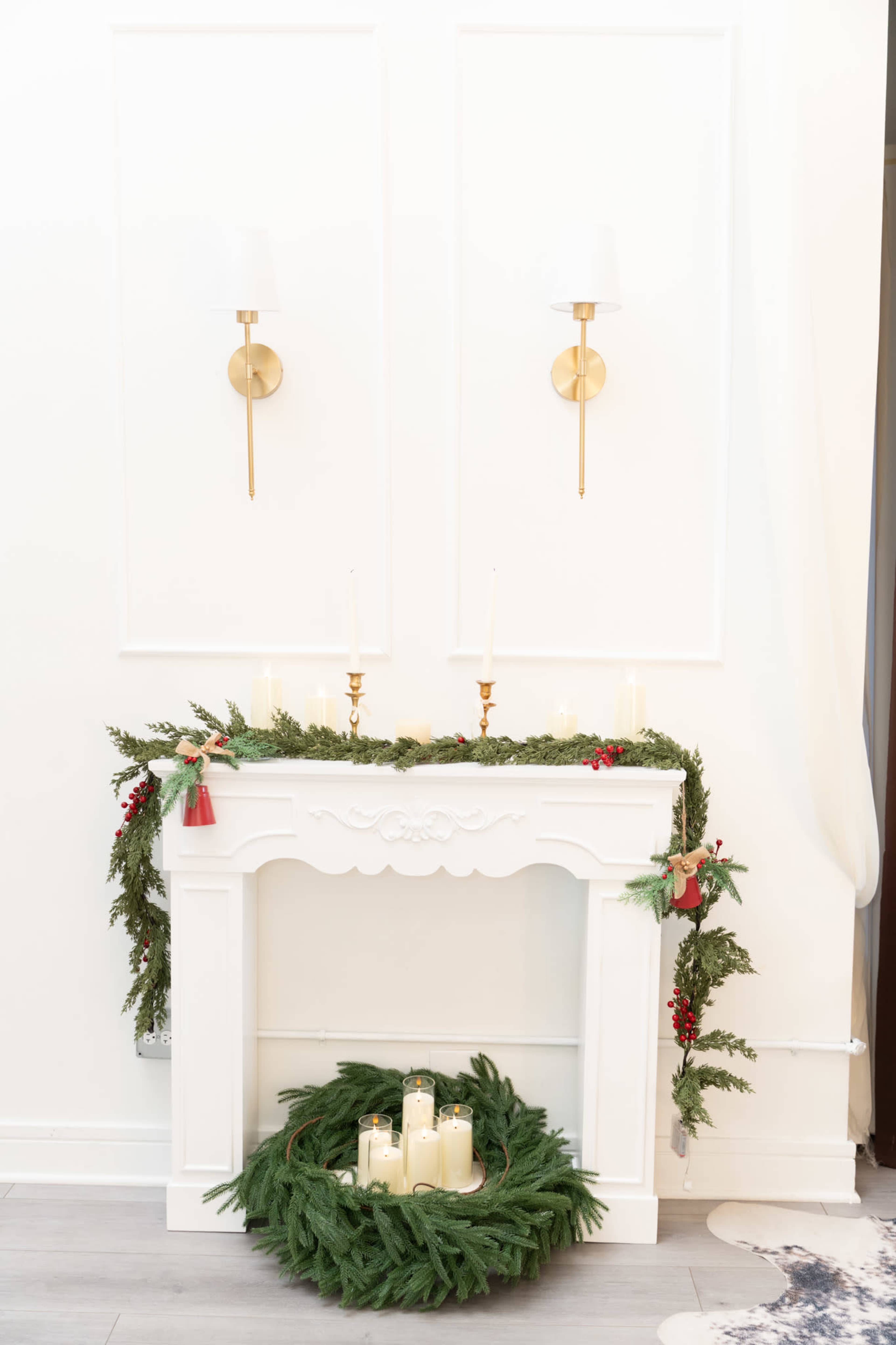 A white mantel decorated with evergreen garland and candles sits against a plain wall, with two gold sconces mounted above.