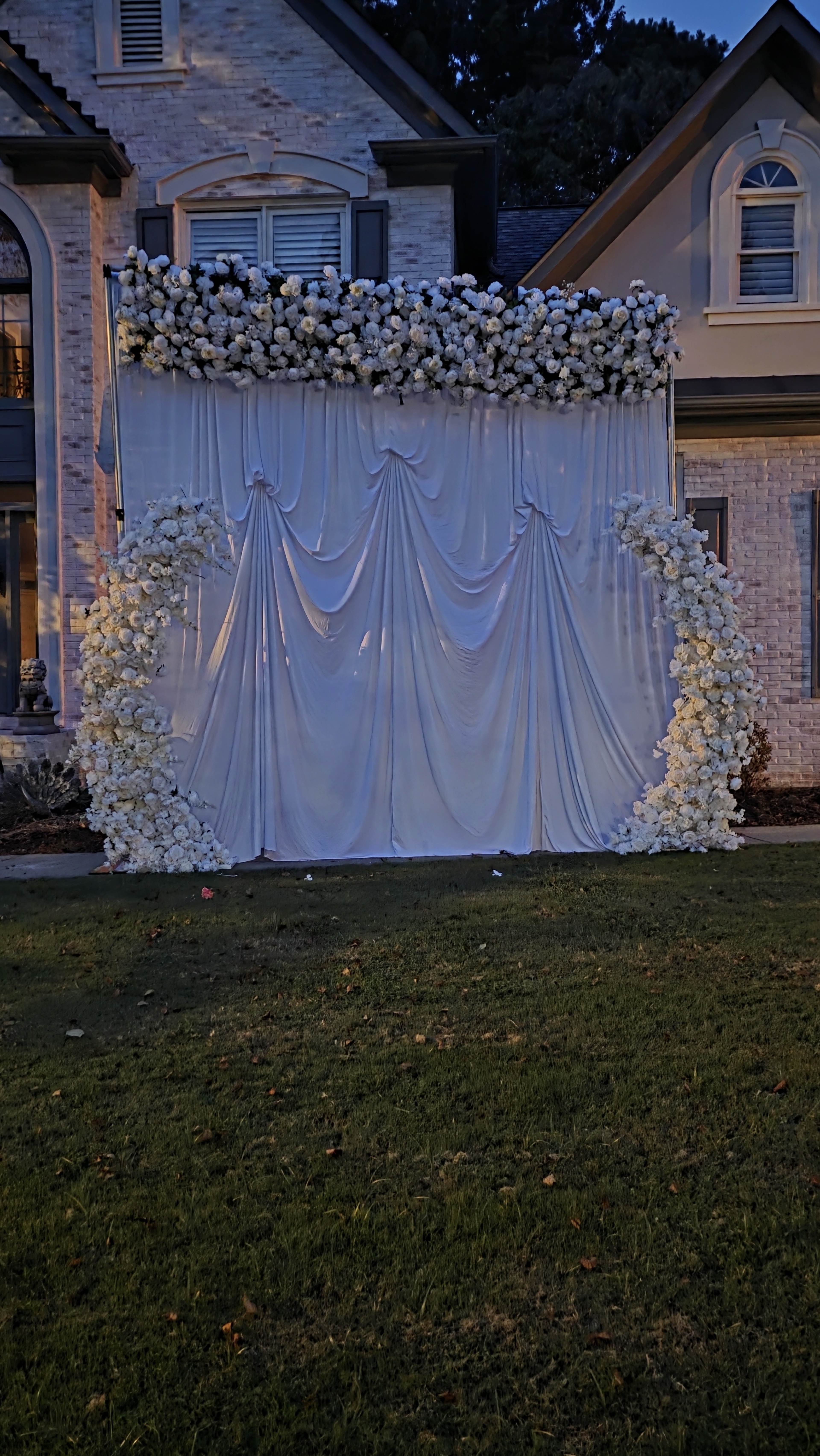 A wedding backdrop featuring white draped fabric and floral arrangements is set up on a lawn in front of a house.
