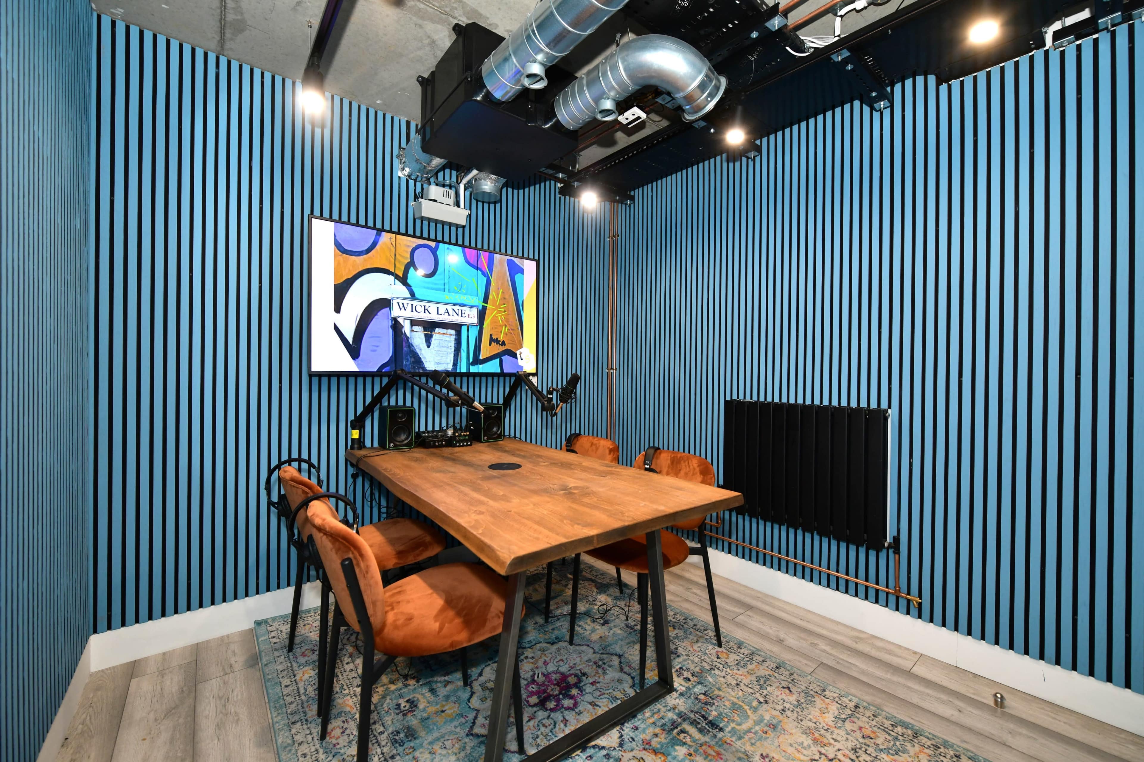 A small meeting room features a wooden table surrounded by four chairs, with a colorful artwork displayed on the wall and blue striped paneling.