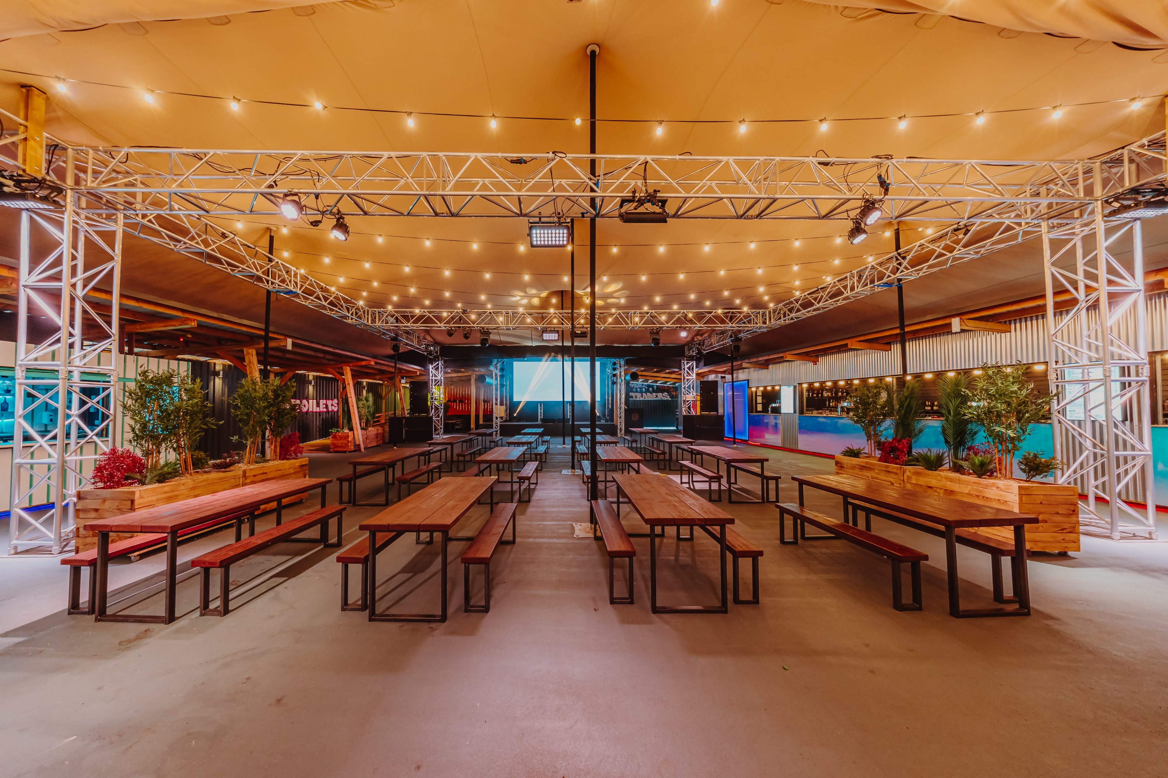 The image shows a spacious indoor venue with long wooden tables and benches, surrounded by string lights and a large screen at the front.