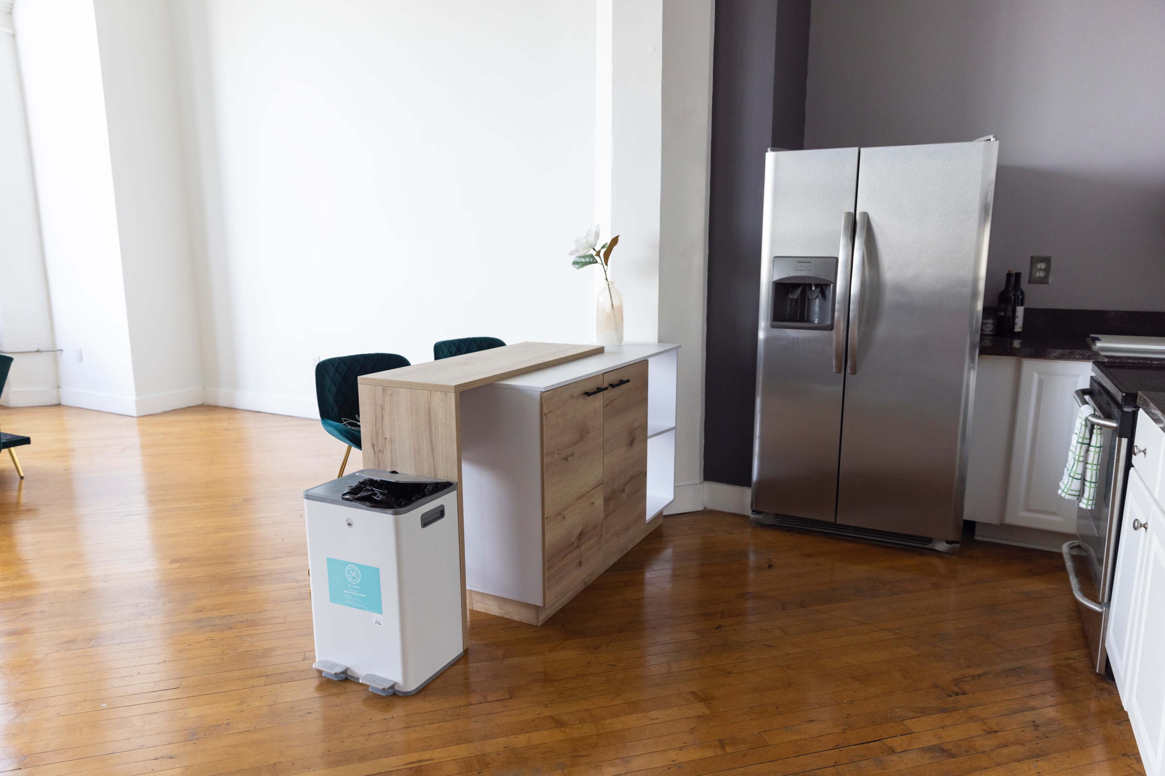 A modern kitchen area with a stainless steel refrigerator, a wooden kitchen island, and a white trash bin on a hardwood floor.