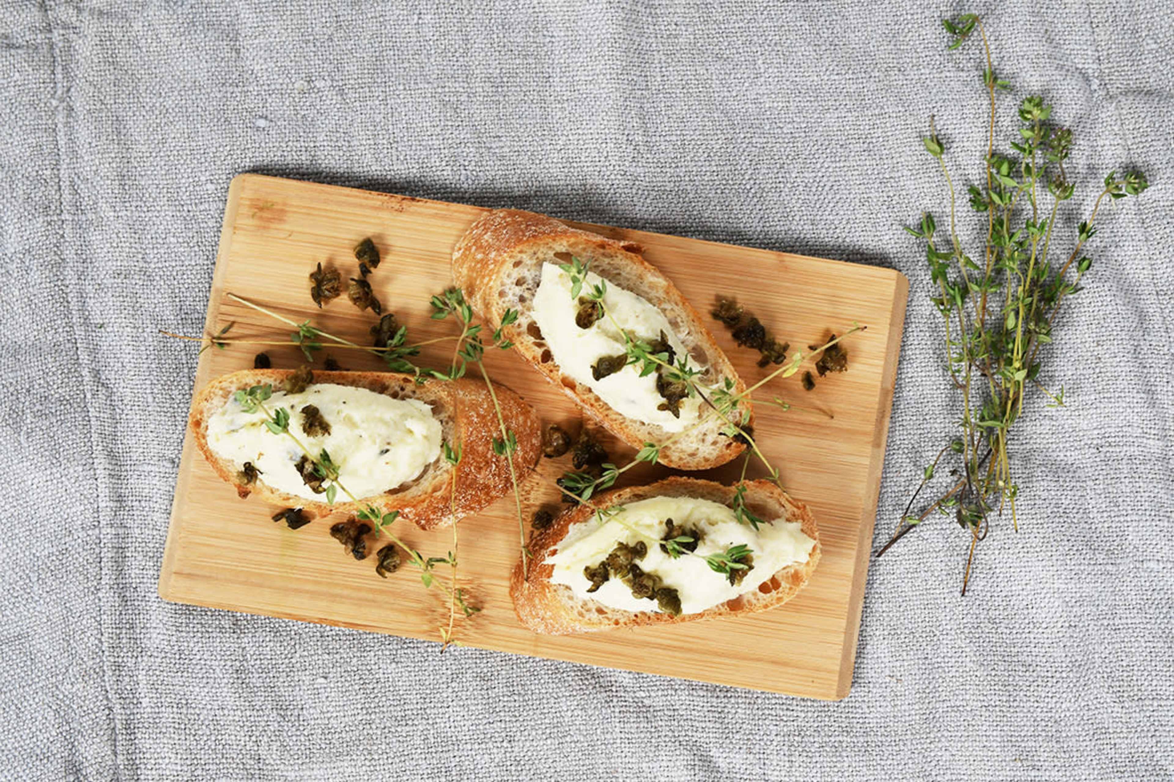 Three slices of toasted bread topped with a creamy spread and garnished with capers and fresh thyme on a wooden cutting board.