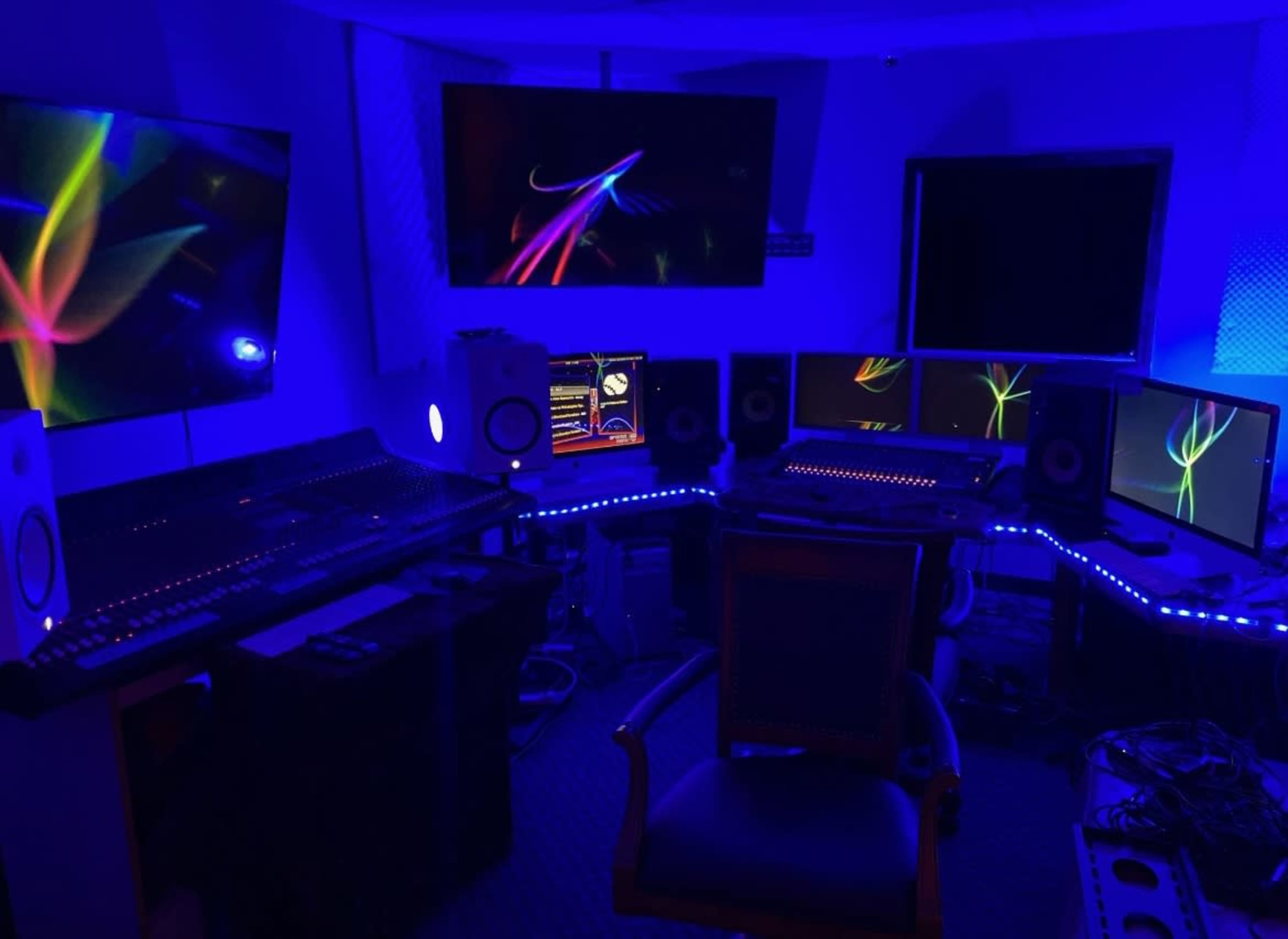 A dimly lit audio studio with multiple computer monitors, a sound mixing console, and ambient blue lighting.