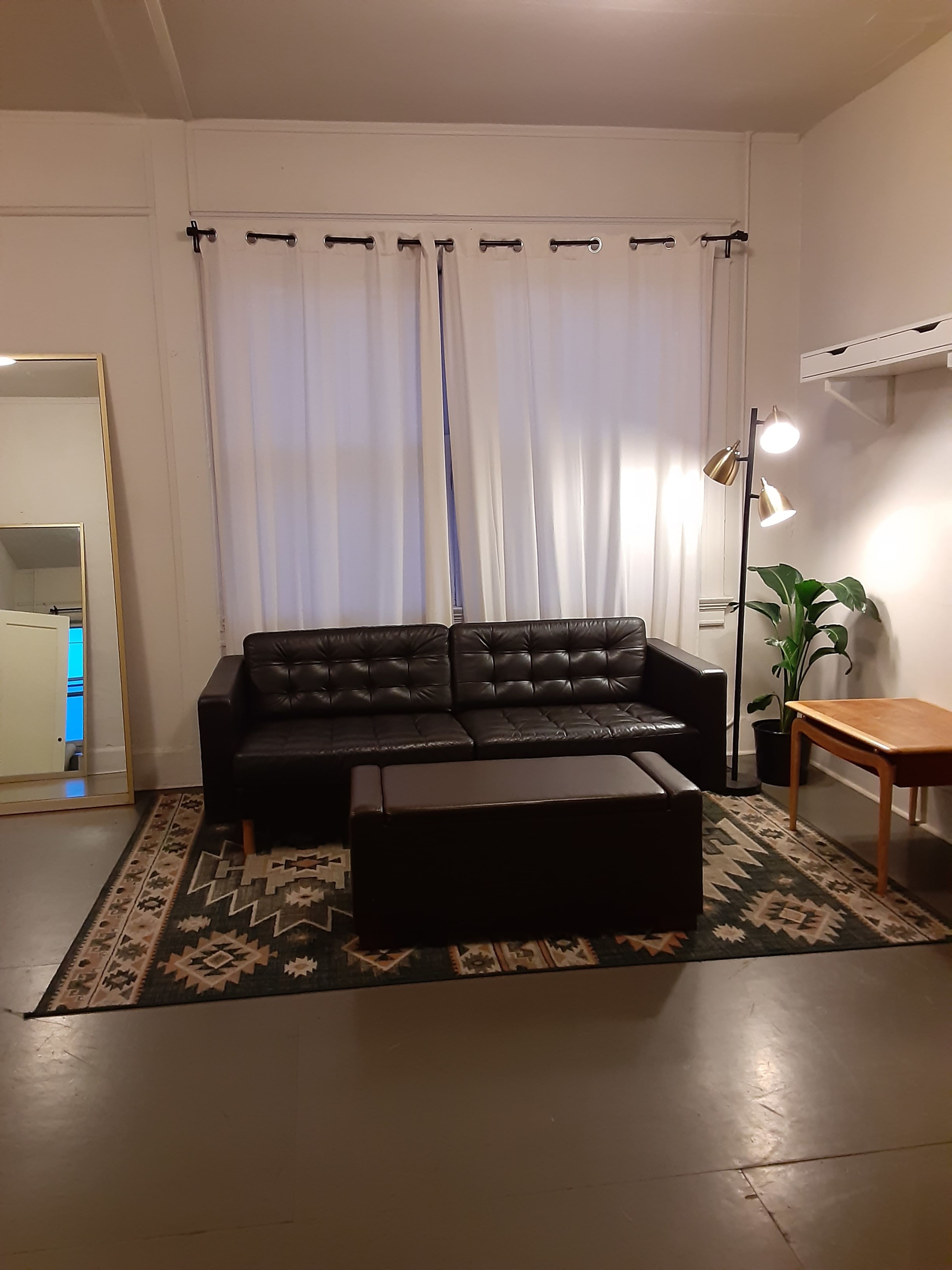 A black leather couch is positioned in front of a large mirror, with white curtains hanging over a window, a floor lamp beside the couch, and a table near a plant on a patterned rug.
