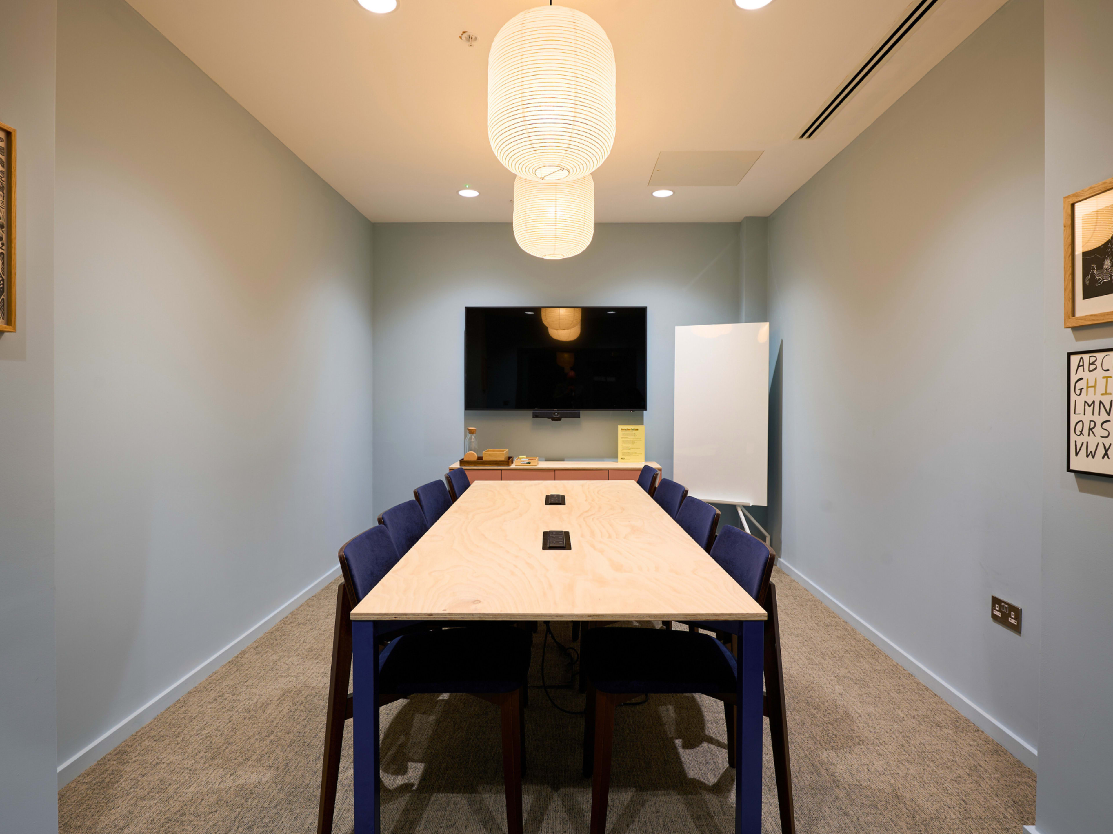 A modern conference room features a long wooden table surrounded by chairs, equipped with a large screen and a whiteboard on the wall.
