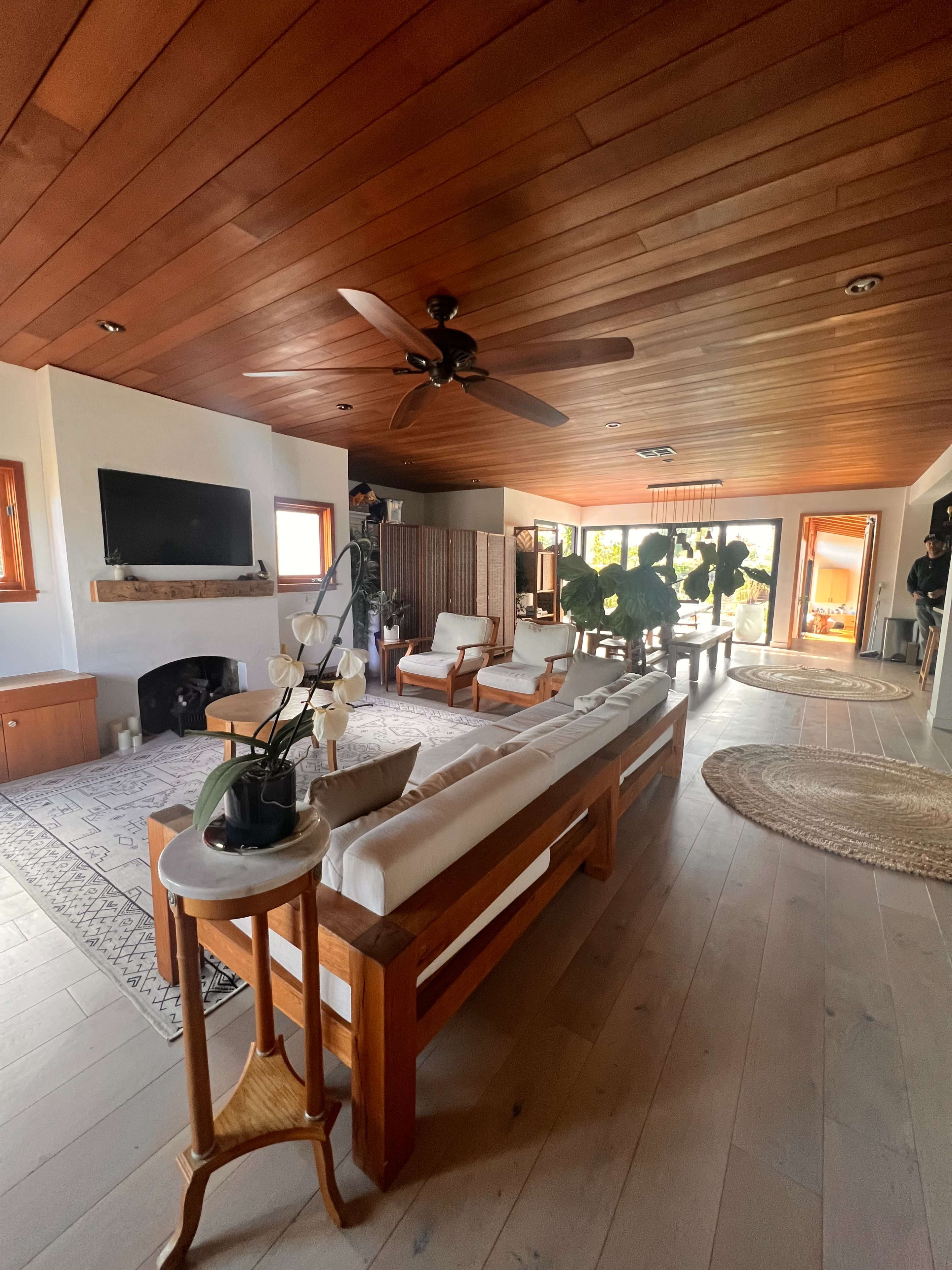 A spacious living room with wooden ceilings, a TV mounted on the wall, and a seating area arranged around a coffee table, leading to an outdoor view.