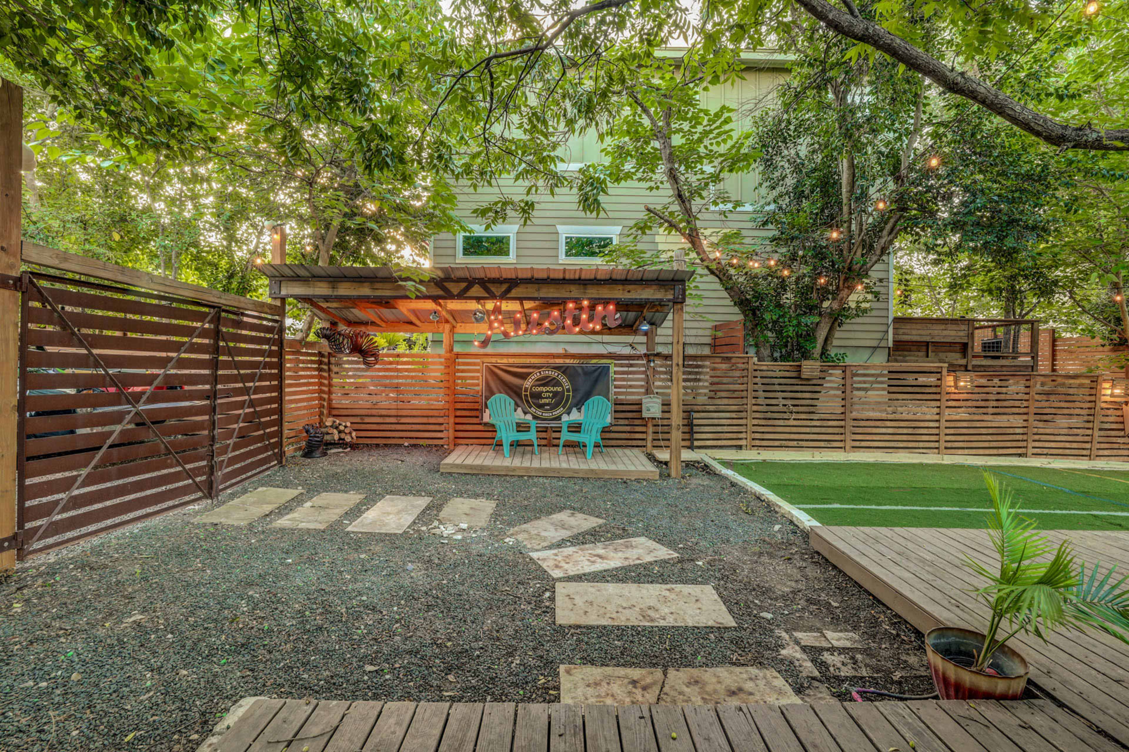 A landscaped backyard features a wooden deck with two blue chairs, a pergola, and a pathway leading to a fenced area.