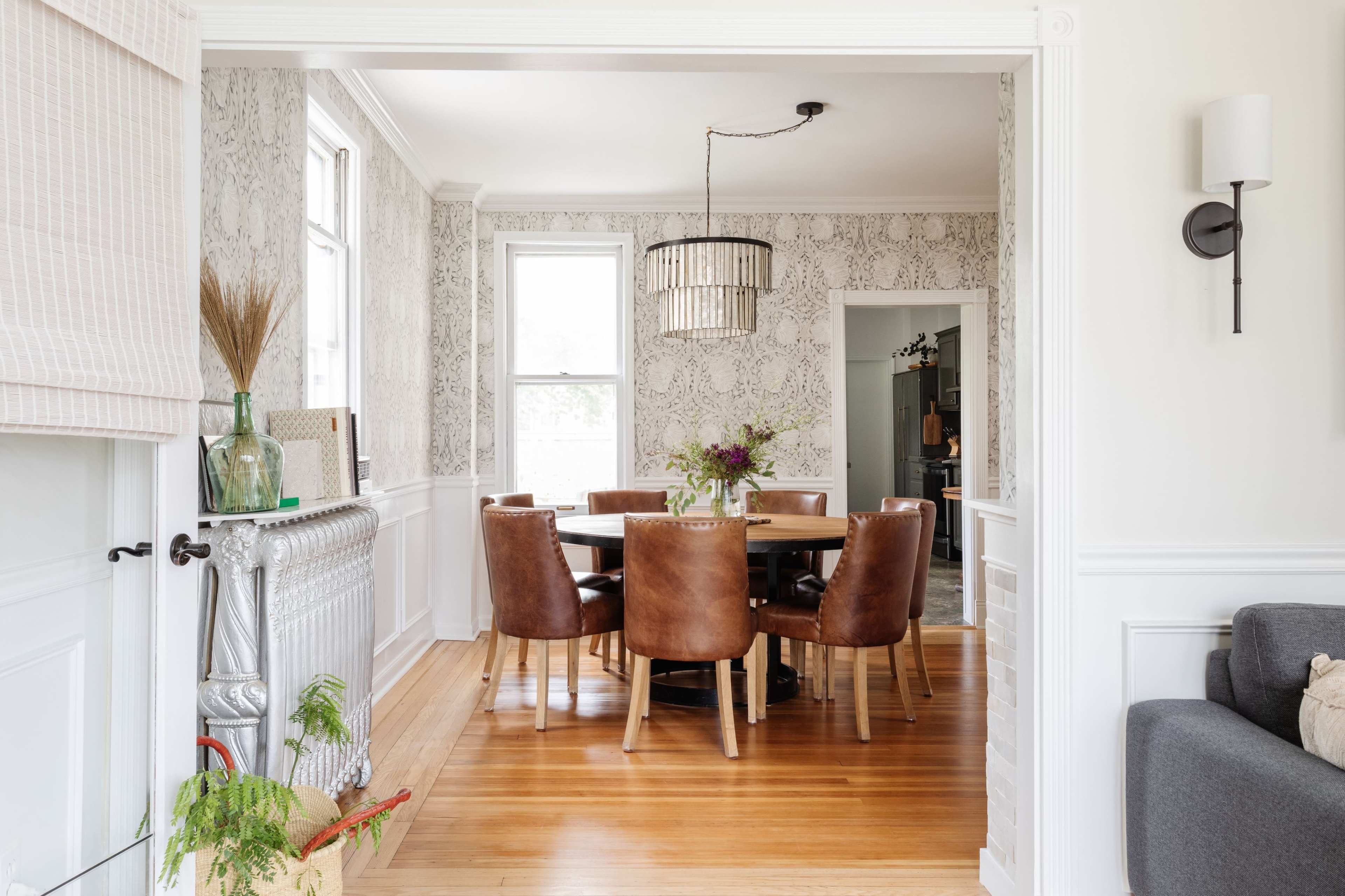 A dining room features a wooden table surrounded by brown leather chairs, with patterned wallpaper and a modern light fixture above.