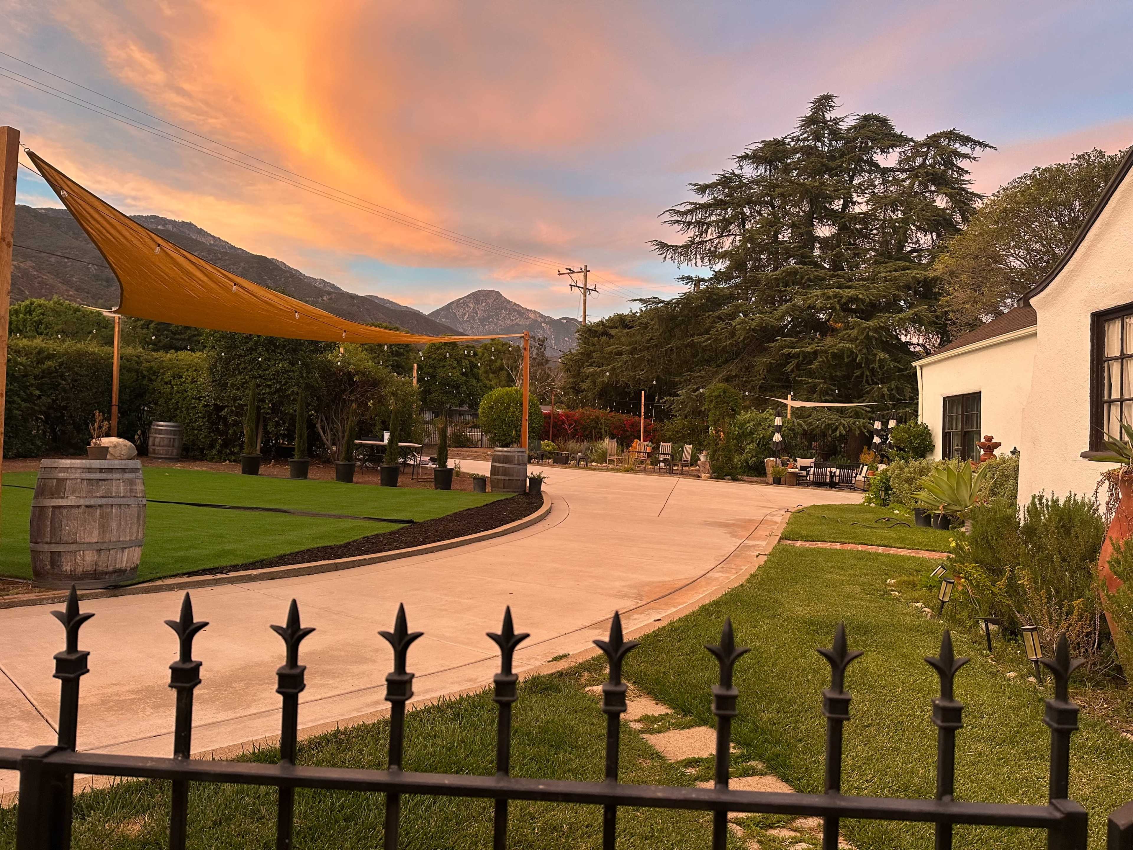 The image depicts a landscaped outdoor area with a curved driveway, a fence in the foreground, and mountains in the background under a colorful sunset sky.