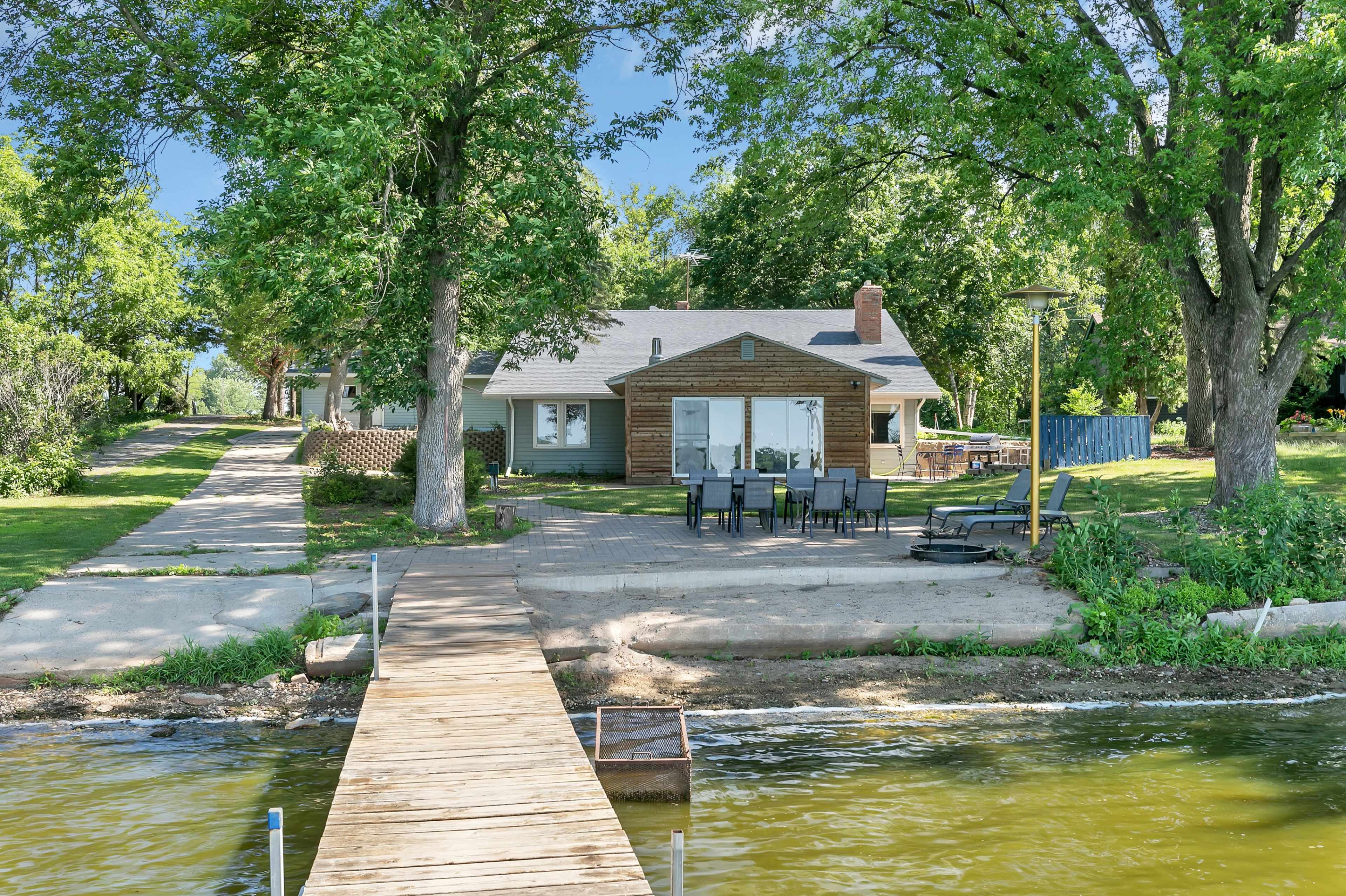 A wooden dock extends over the water toward a house set back from the shore, surrounded by trees and a patio area with outdoor seating.