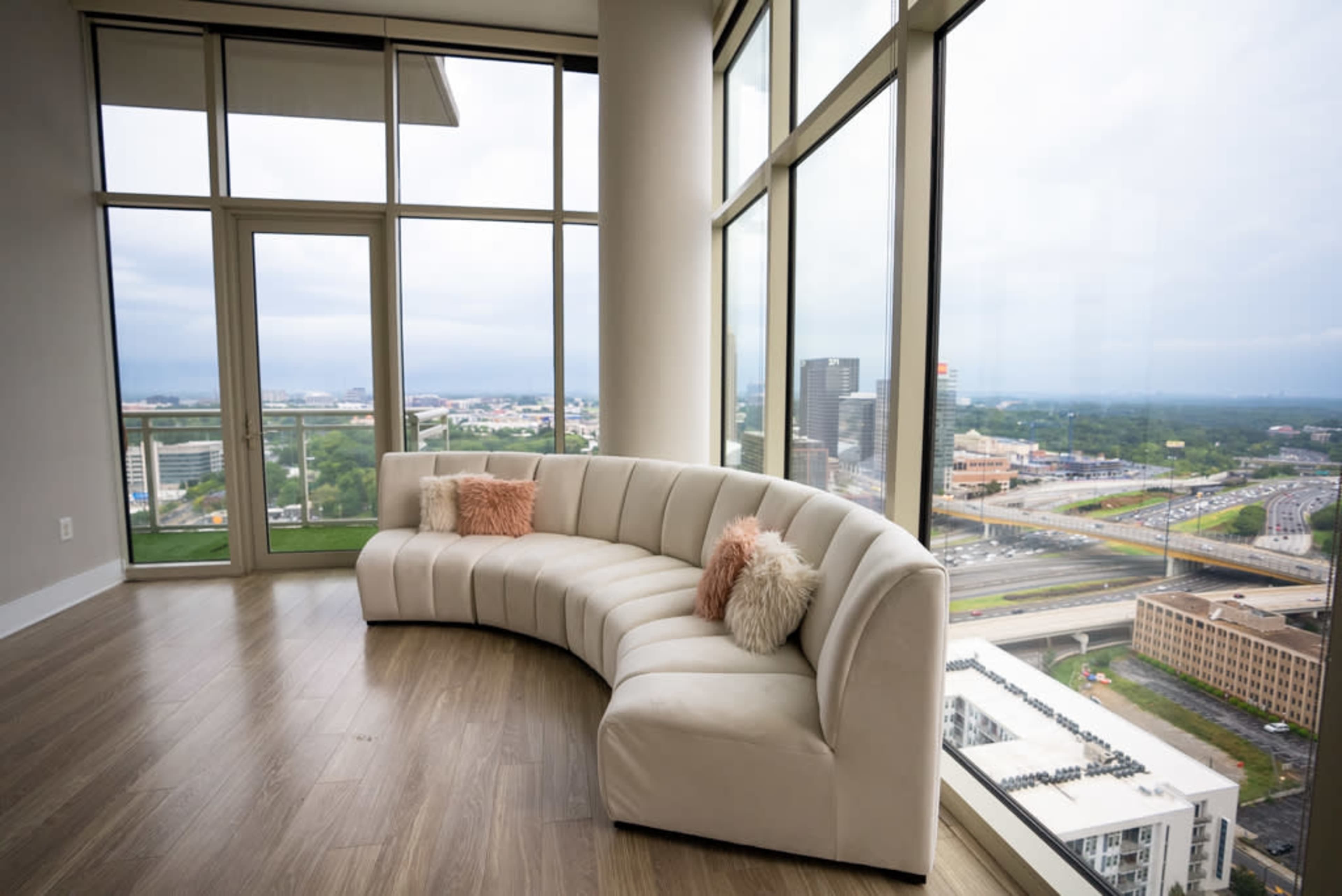 A curved white sofa is positioned in a bright room with large windows overlooking a cityscape.