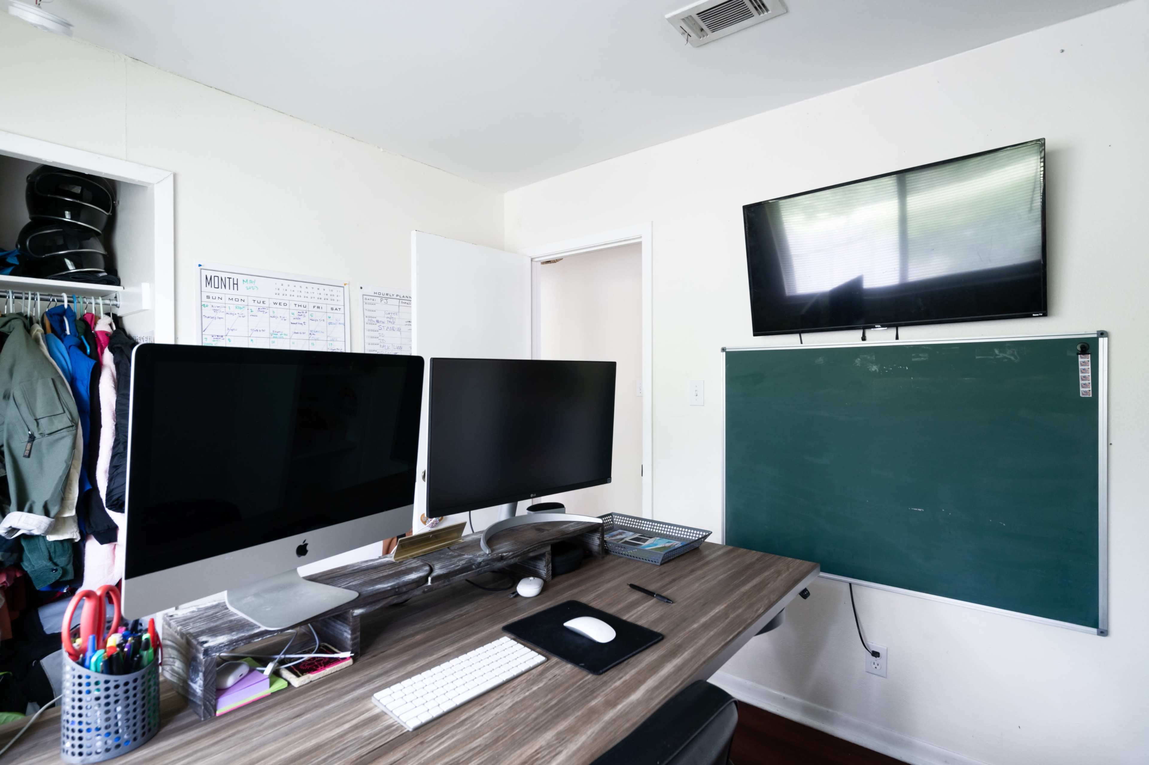 The image shows a home office setup with two large computer monitors, a desk cluttered with office supplies, a green chalkboard mounted on the wall, and a television above it.
