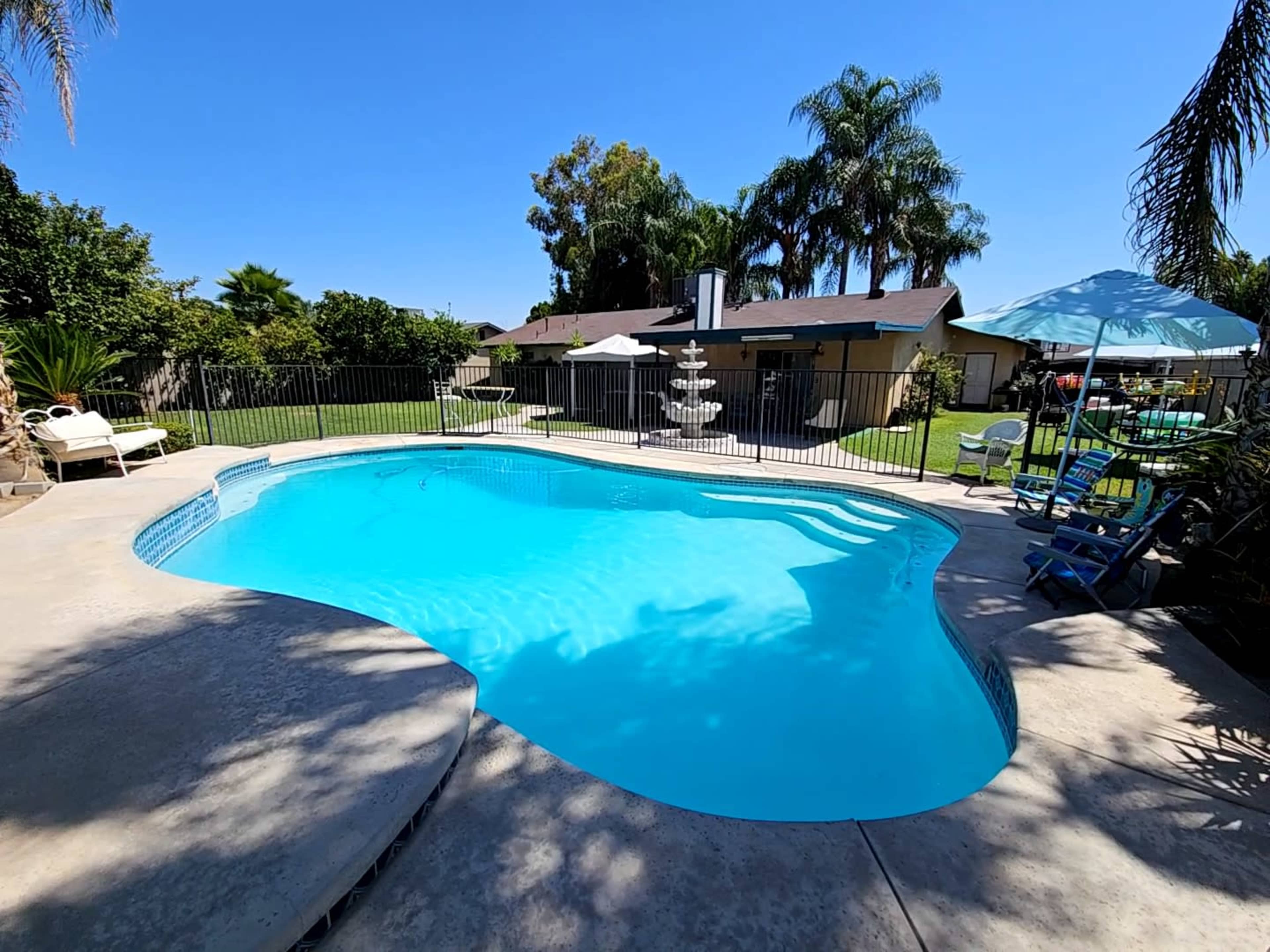 A clear swimming pool is surrounded by a concrete patio and a lush green backyard with palm trees and outdoor furniture.