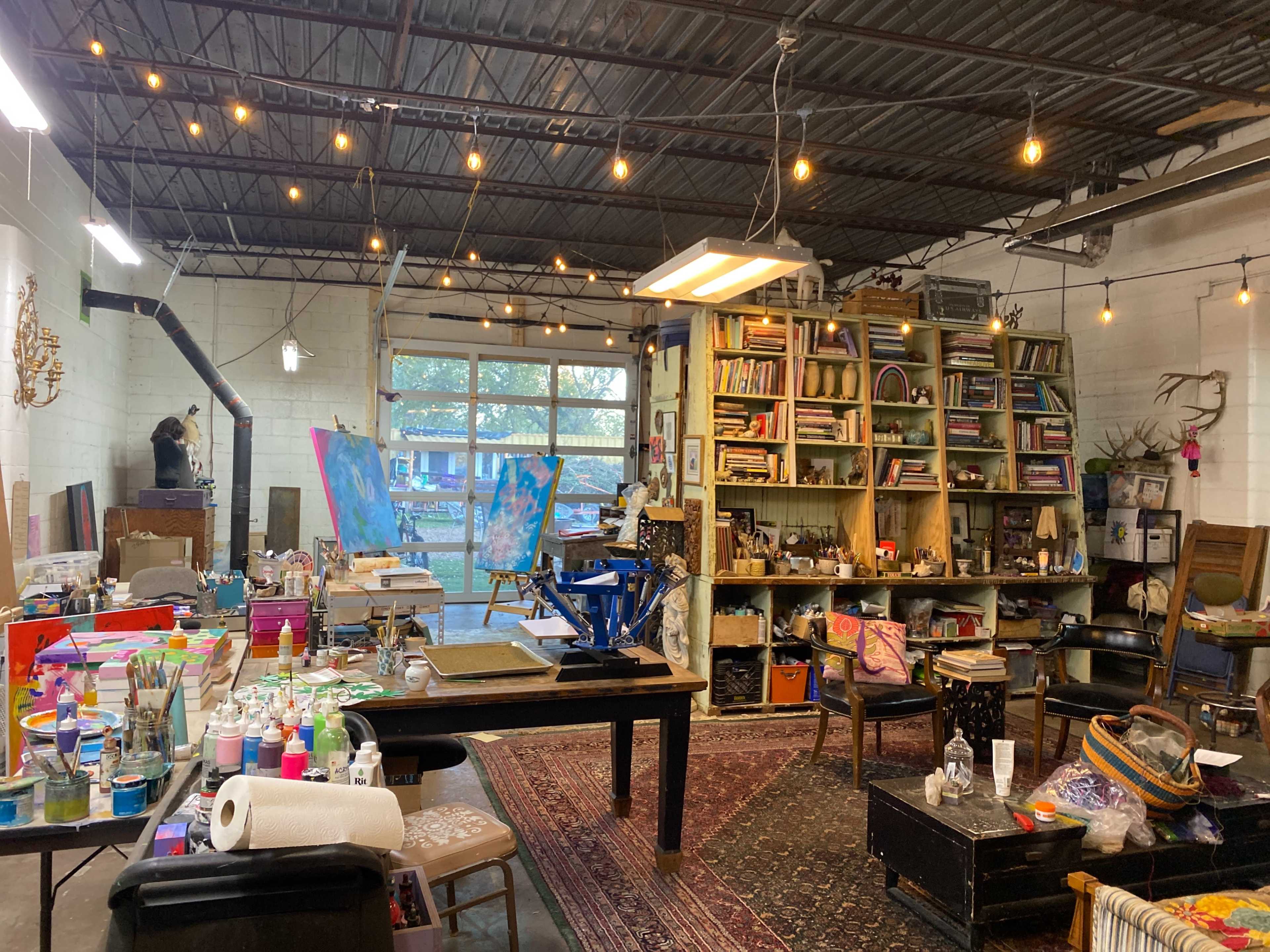 The image shows a cluttered art studio featuring a large shelving unit filled with materials, various art supplies on tables, and canvases displayed in the background.