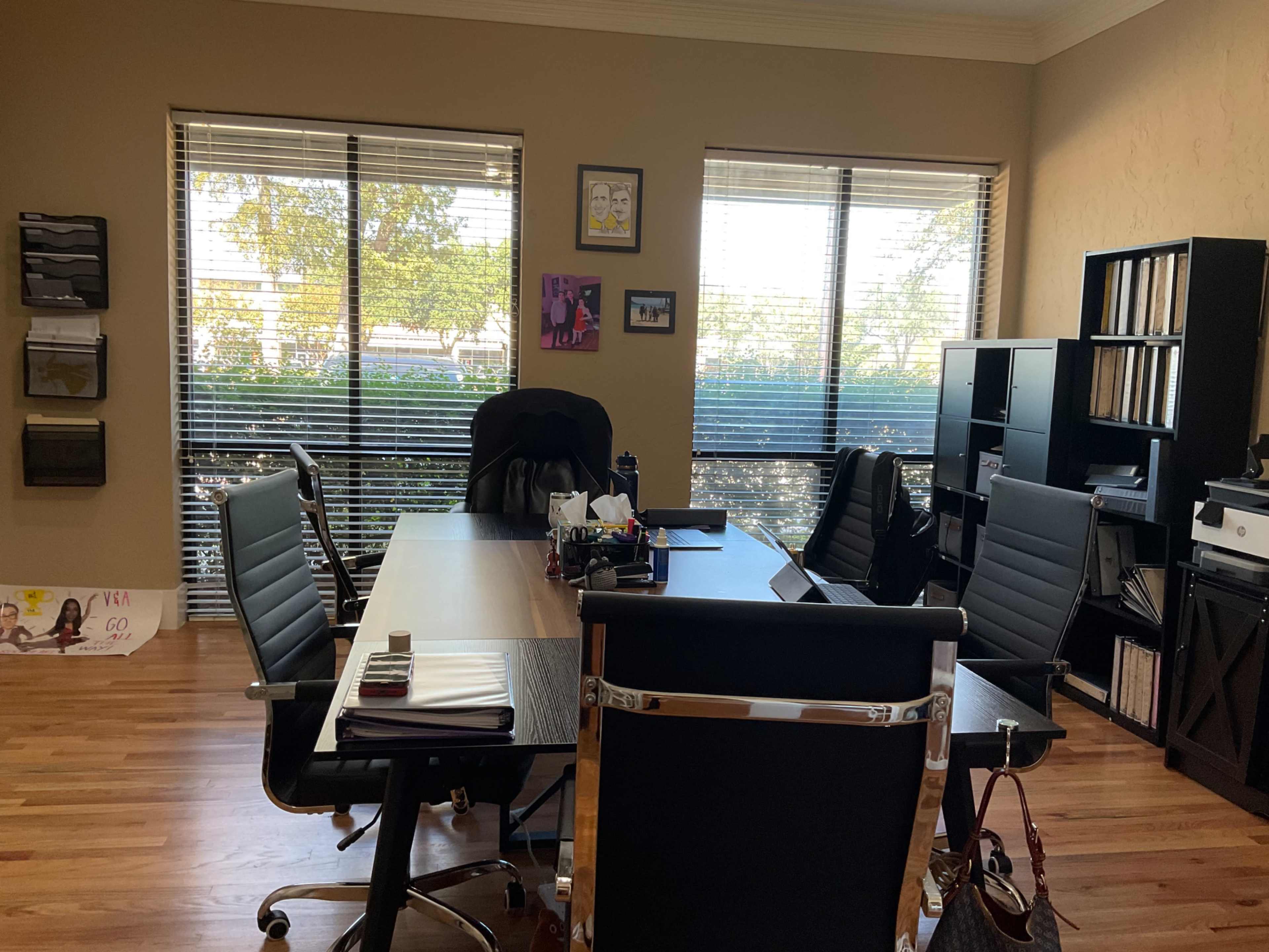 The image shows a modern office space featuring a central desk, several black office chairs, and large windows with natural light illuminating the room.