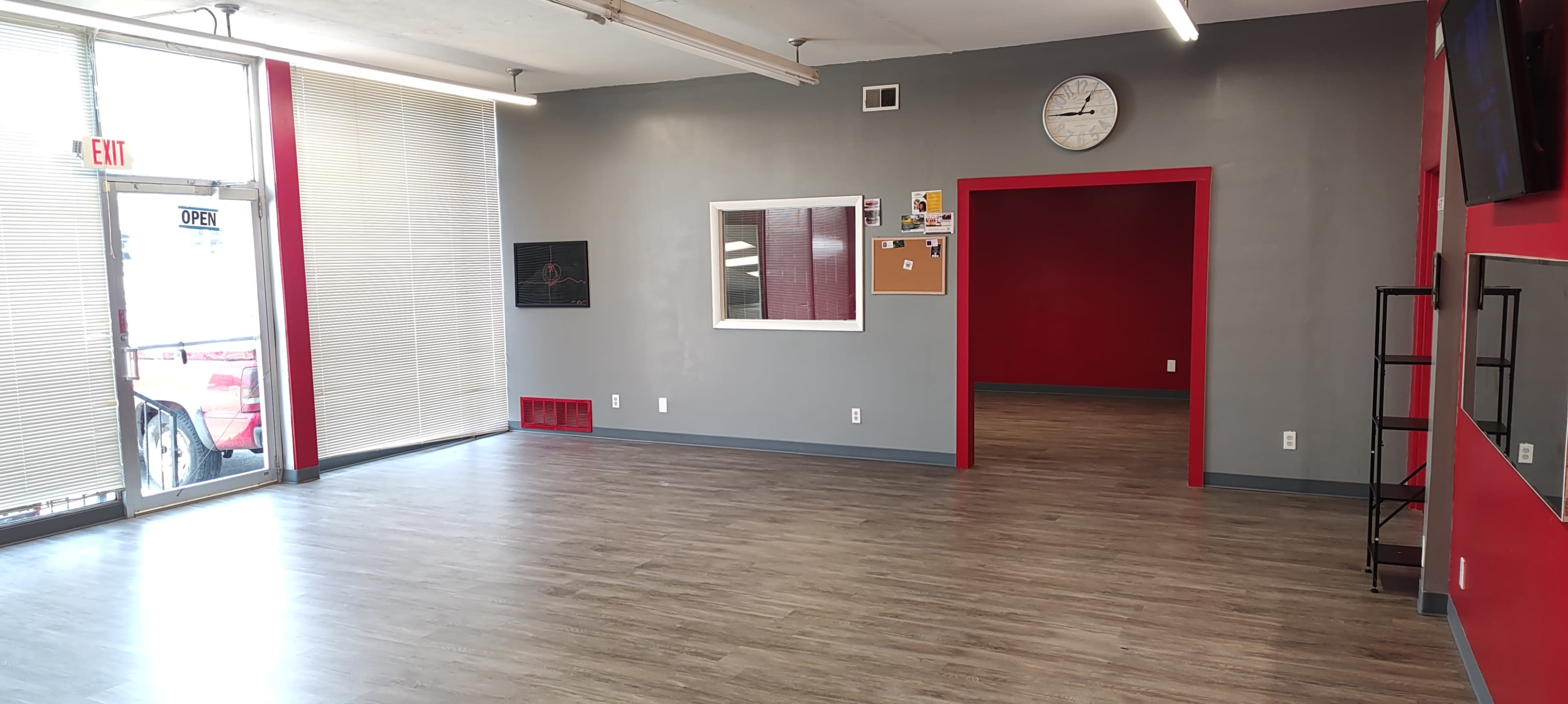The image shows an empty room with gray walls, wooden flooring, and a large window displaying an entrance.
