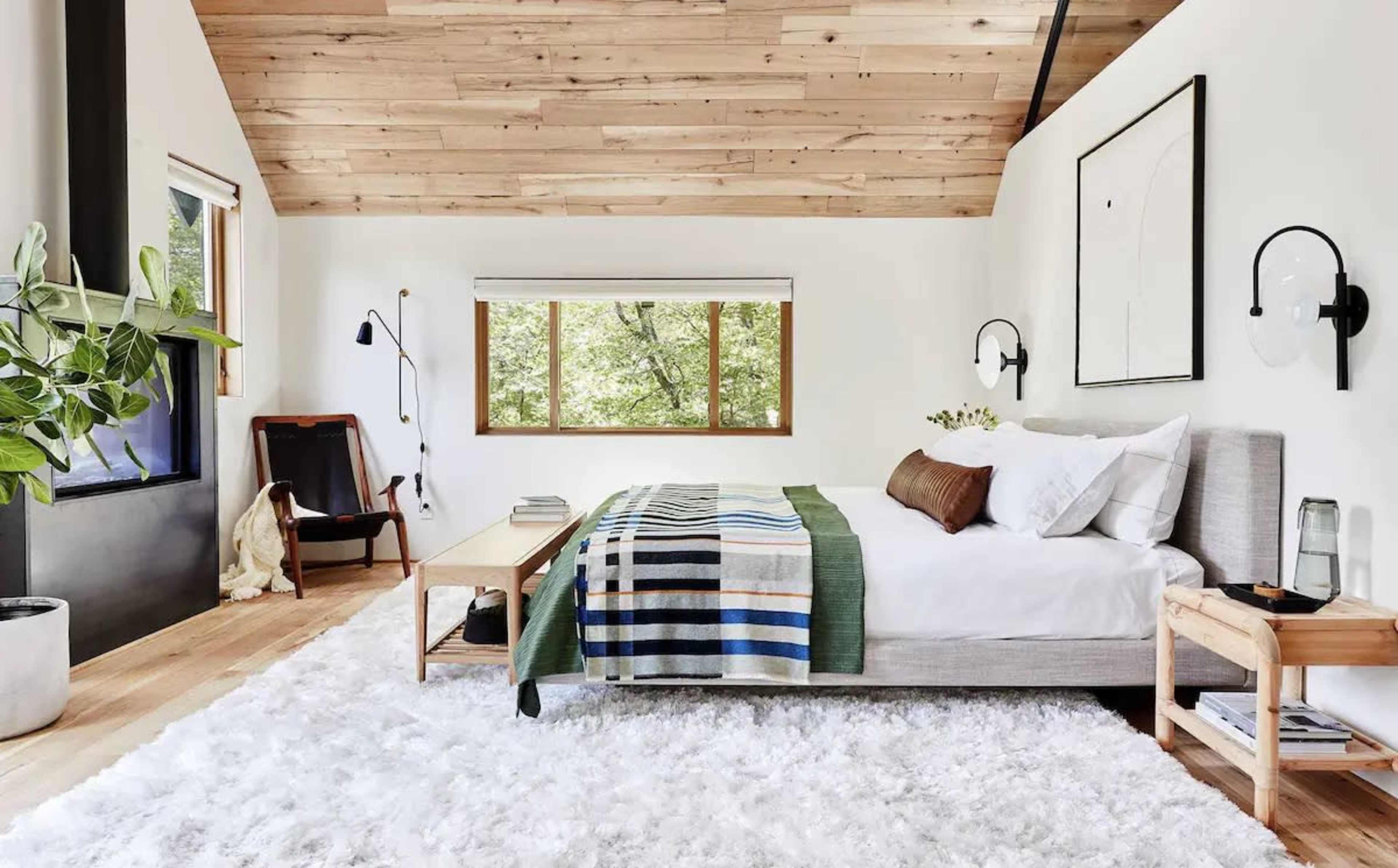 A modern bedroom with a bed, a wooden ceiling, large windows, and a cozy rug.