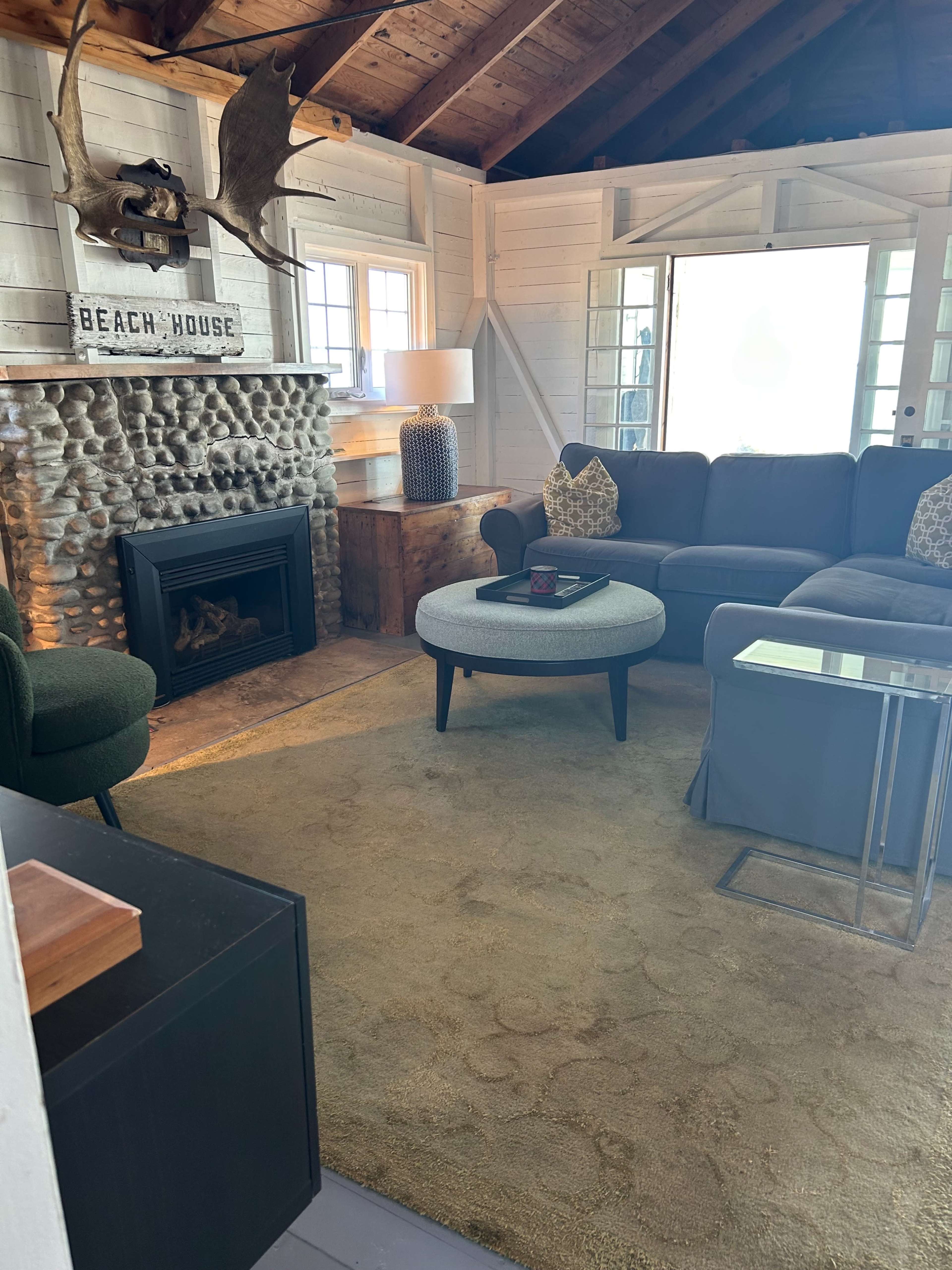 The image shows a cozy living room featuring a stone fireplace, a round ottoman, a sectional sofa, and large windows allowing natural light.