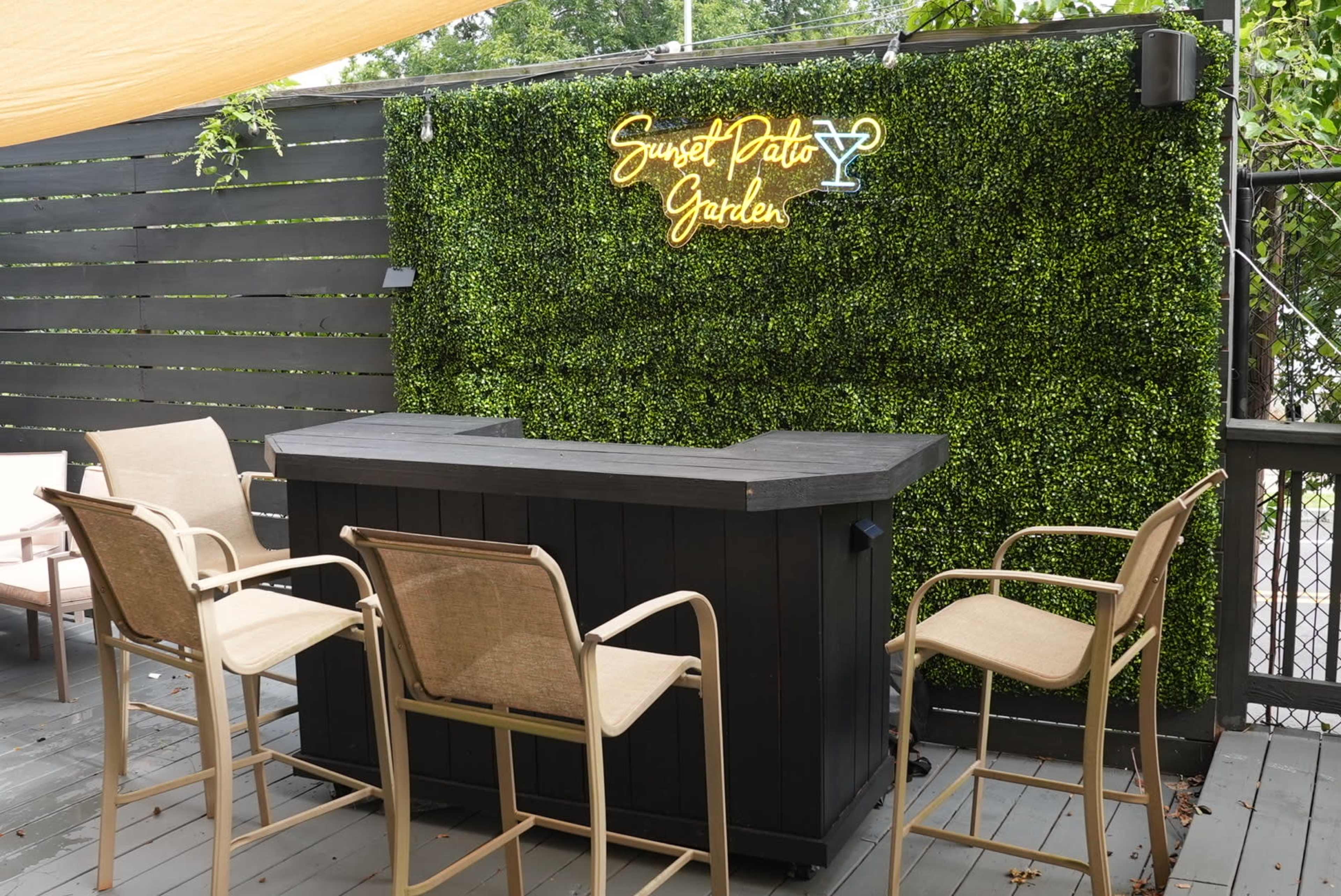 The image shows a bar area with four tan chairs in front of a black wooden bar and a green foliage backdrop featuring a neon sign that reads "Sunset Patio Garden."