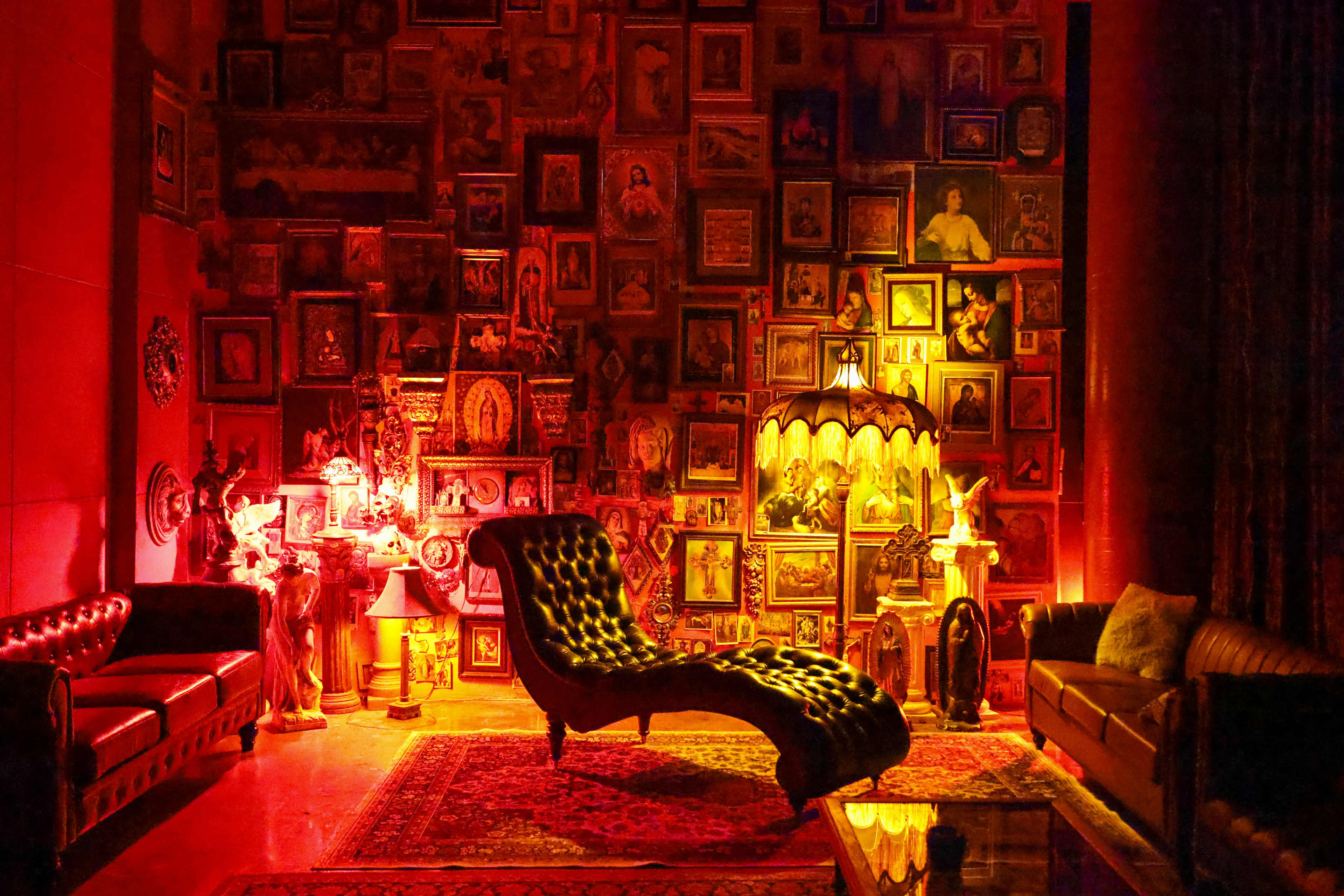 The room features a vintage chaise lounge and two sofas against a wall filled with framed artwork, illuminated by warm lighting from a decorative lamp.