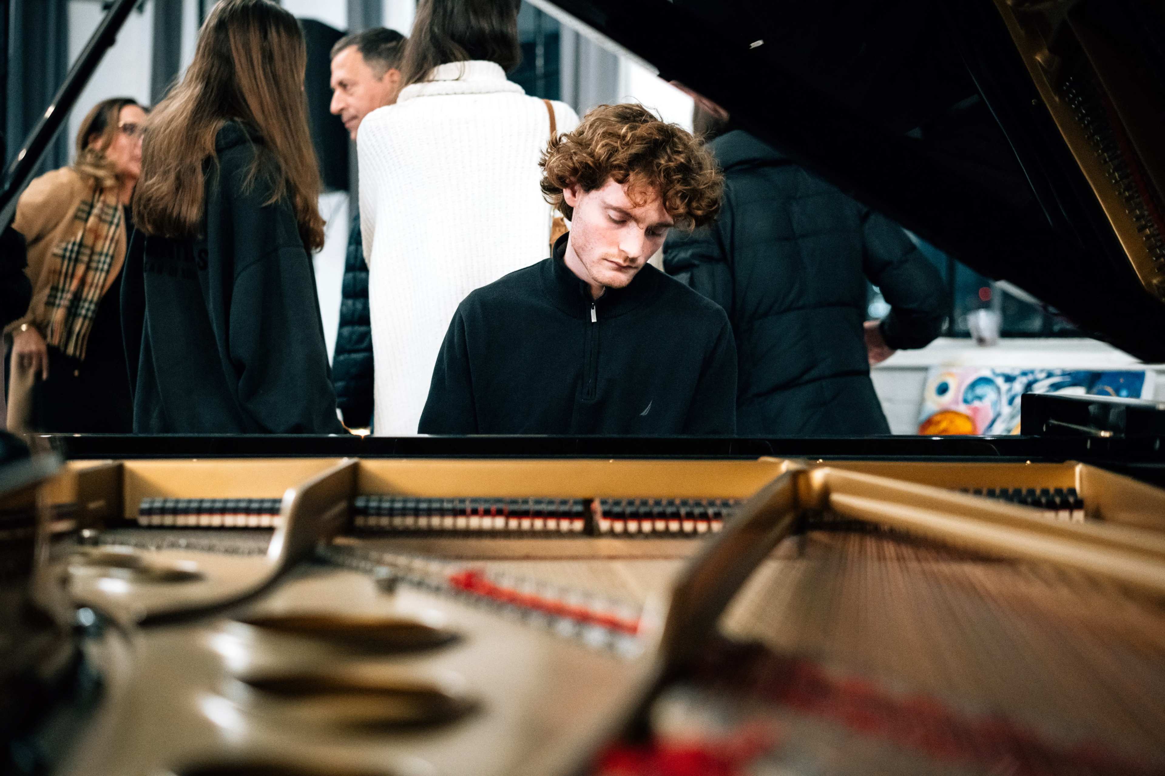 A young man plays a piano in the foreground while people converse in the background.