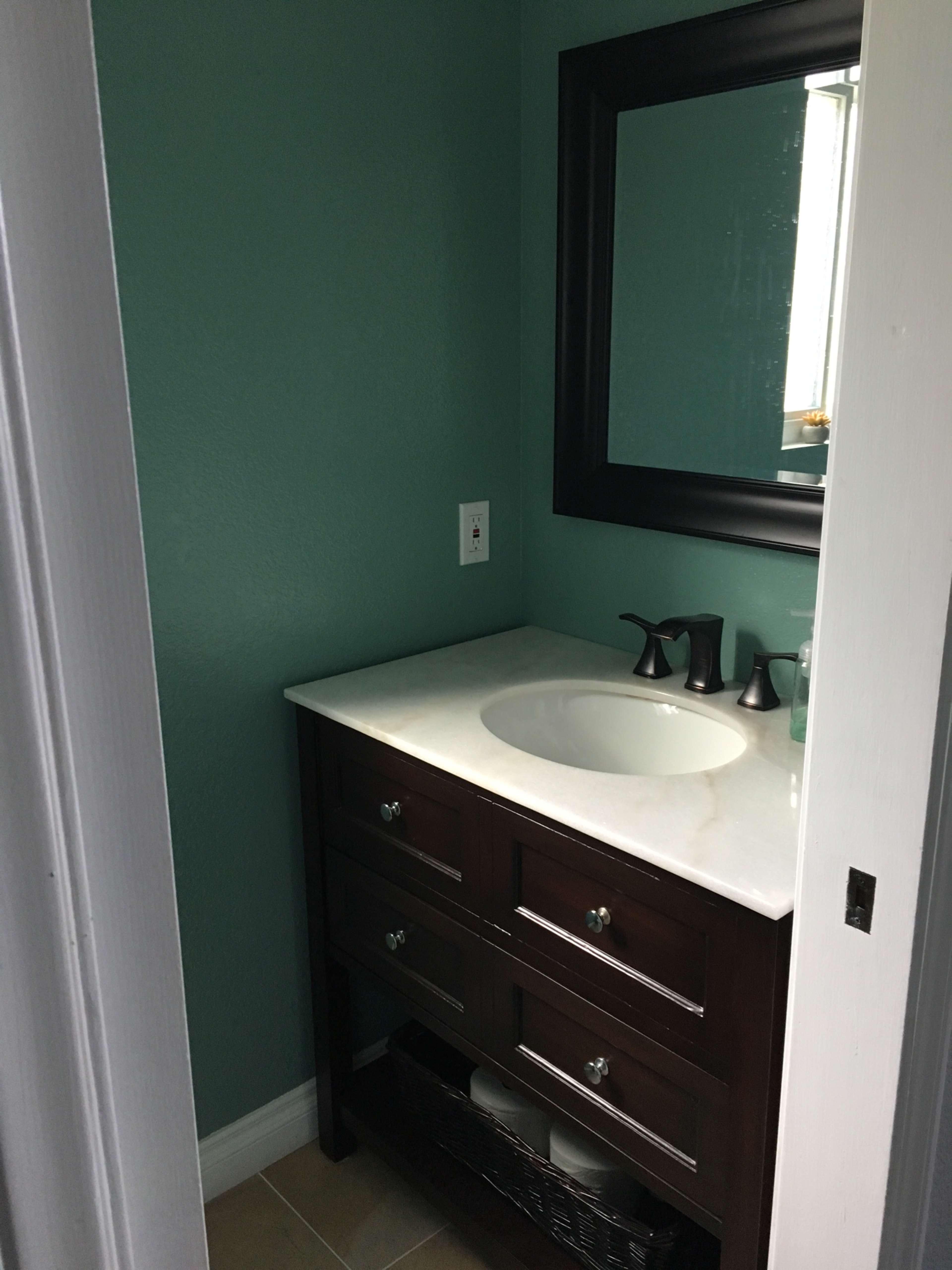 A small bathroom with a dark wood vanity, a white countertop, and a mirror framed in black against a teal wall.