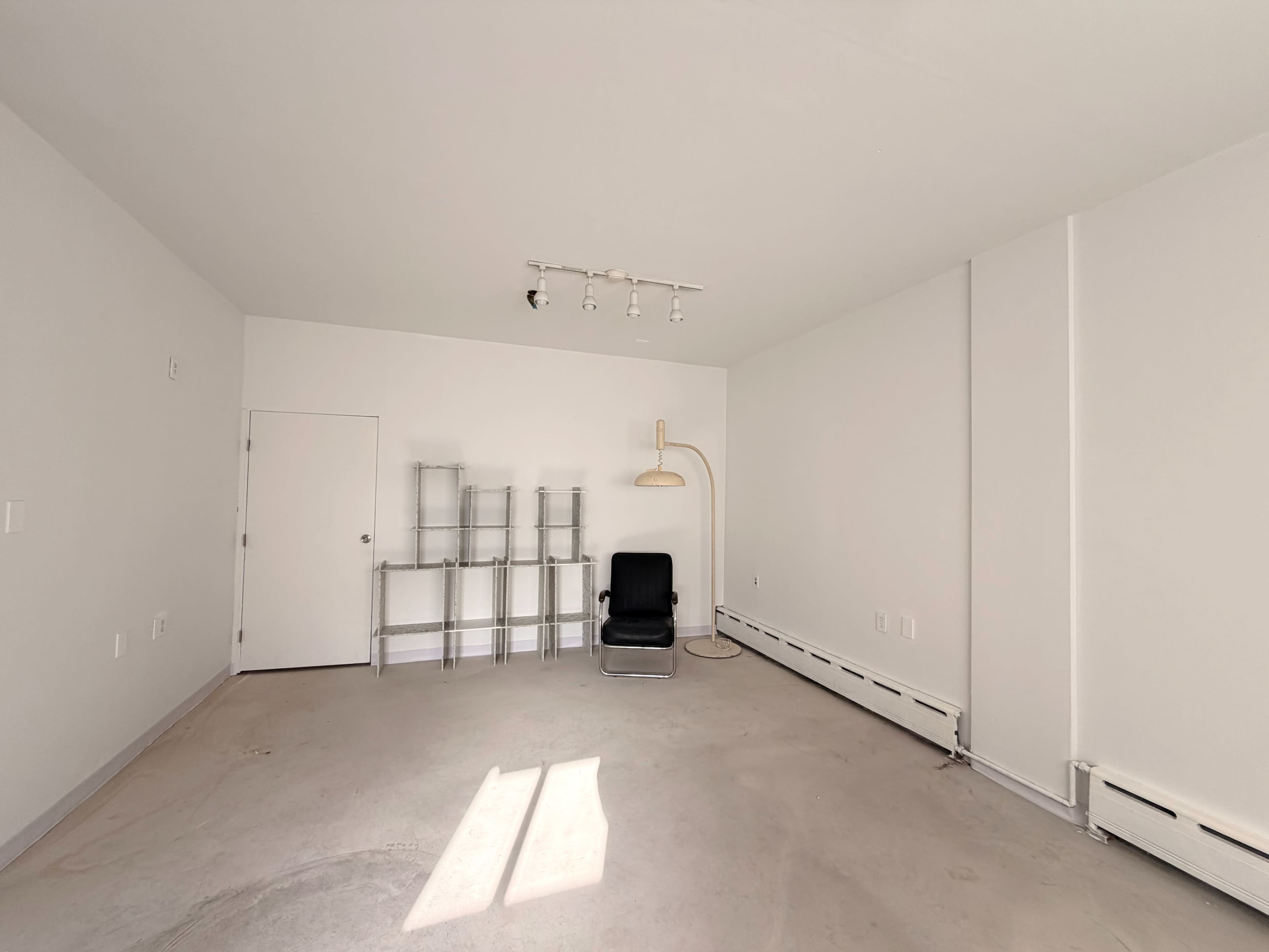 The image shows an empty, well-lit room with a chair, a floor lamp, and a metallic shelf unit against one wall.