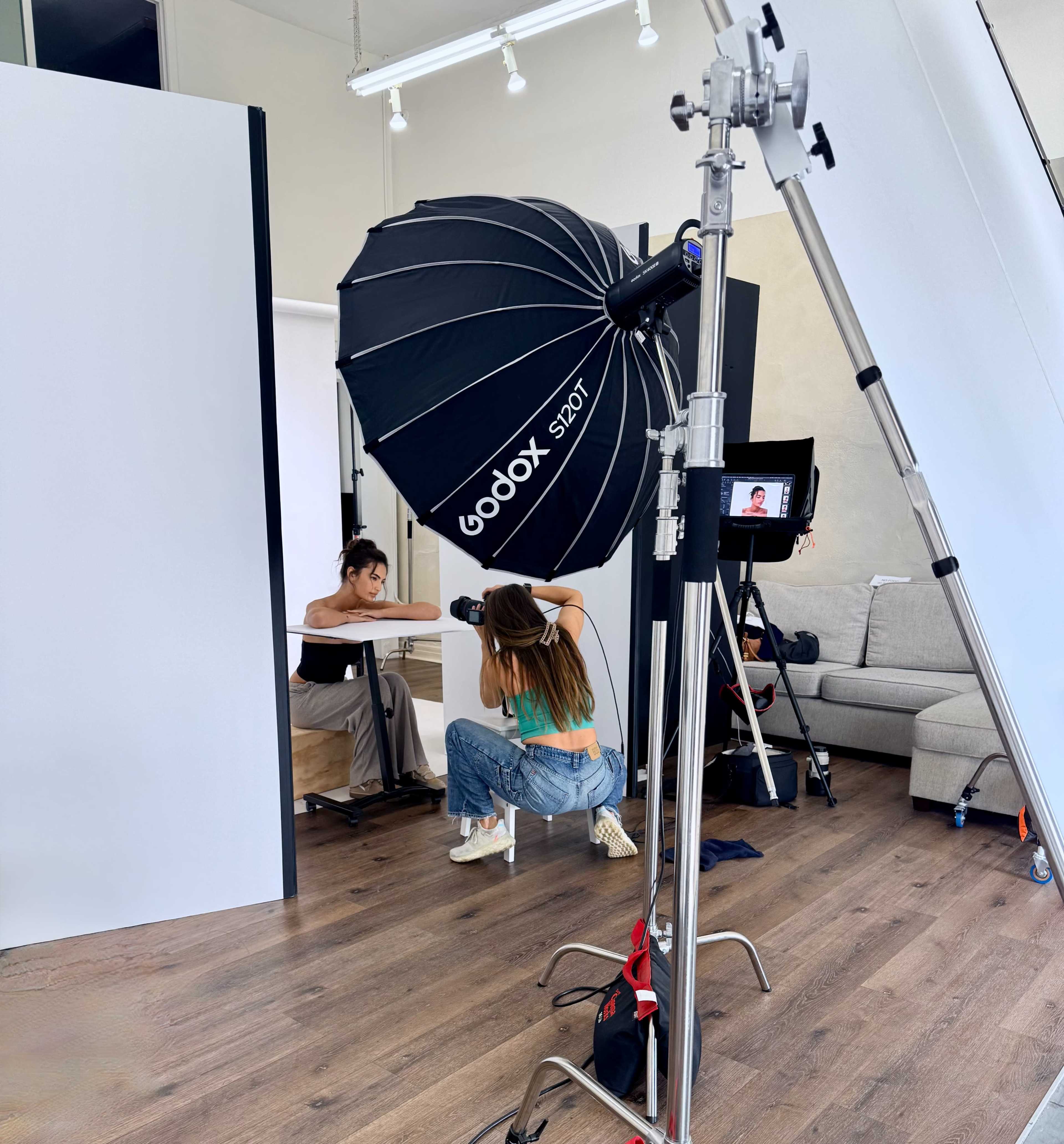 A photography studio setup features a photographer positioning a light umbrella while a model sits at a table nearby.