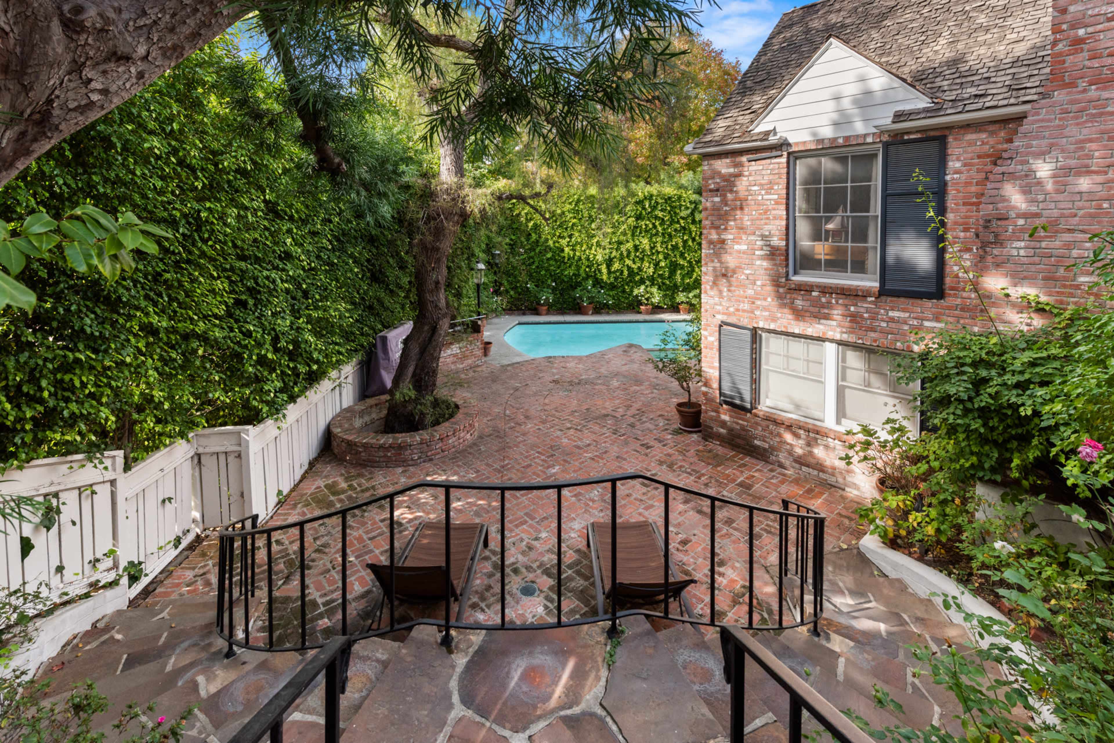 A brick patio with two lounge chairs overlooks a swimming pool surrounded by lush greenery and a house with white and brick facades.