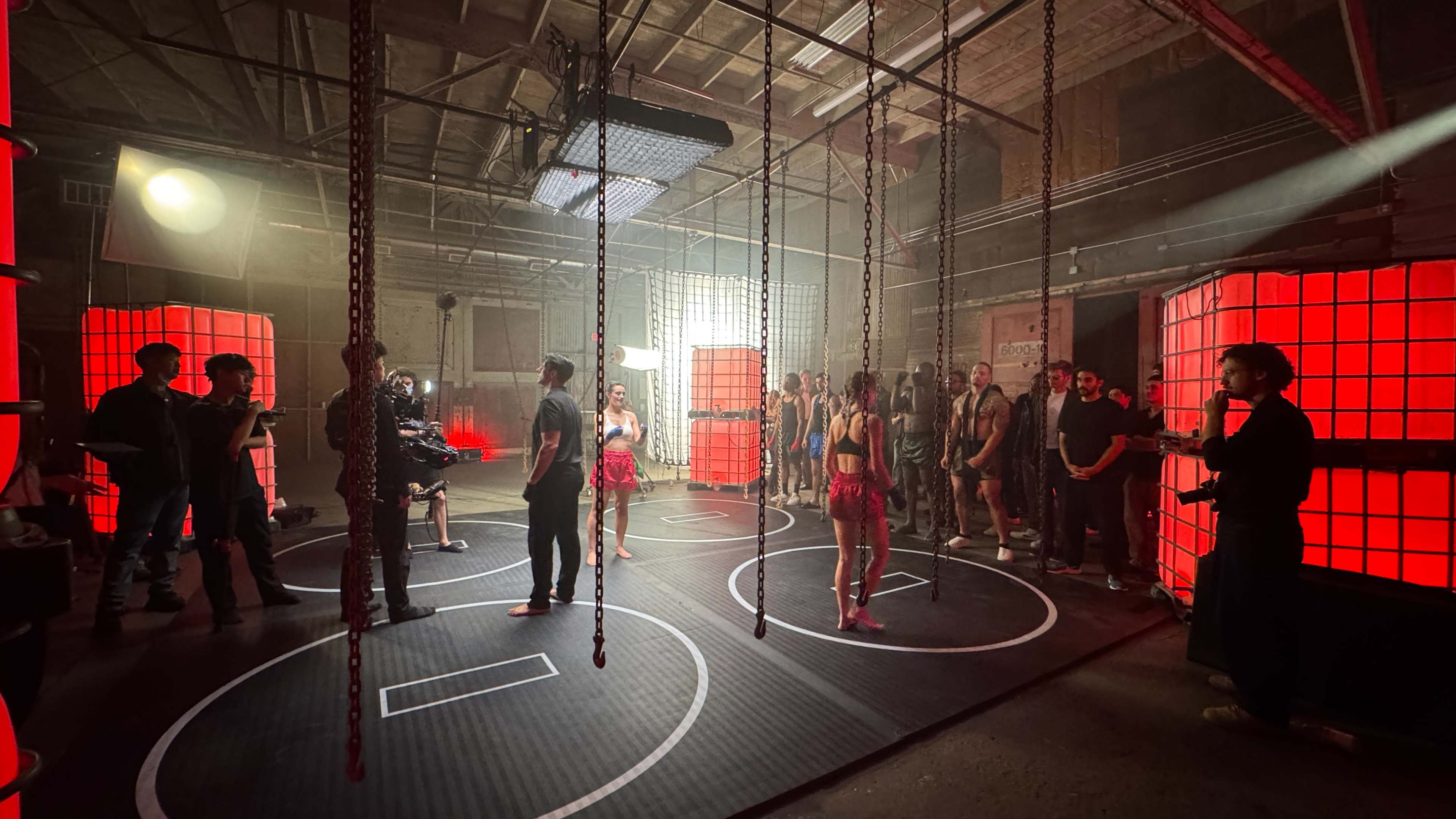 A group of people is gathered in a dimly lit warehouse, surrounded by red-lit boxes and suspended chains, while a camera crew captures the scene.