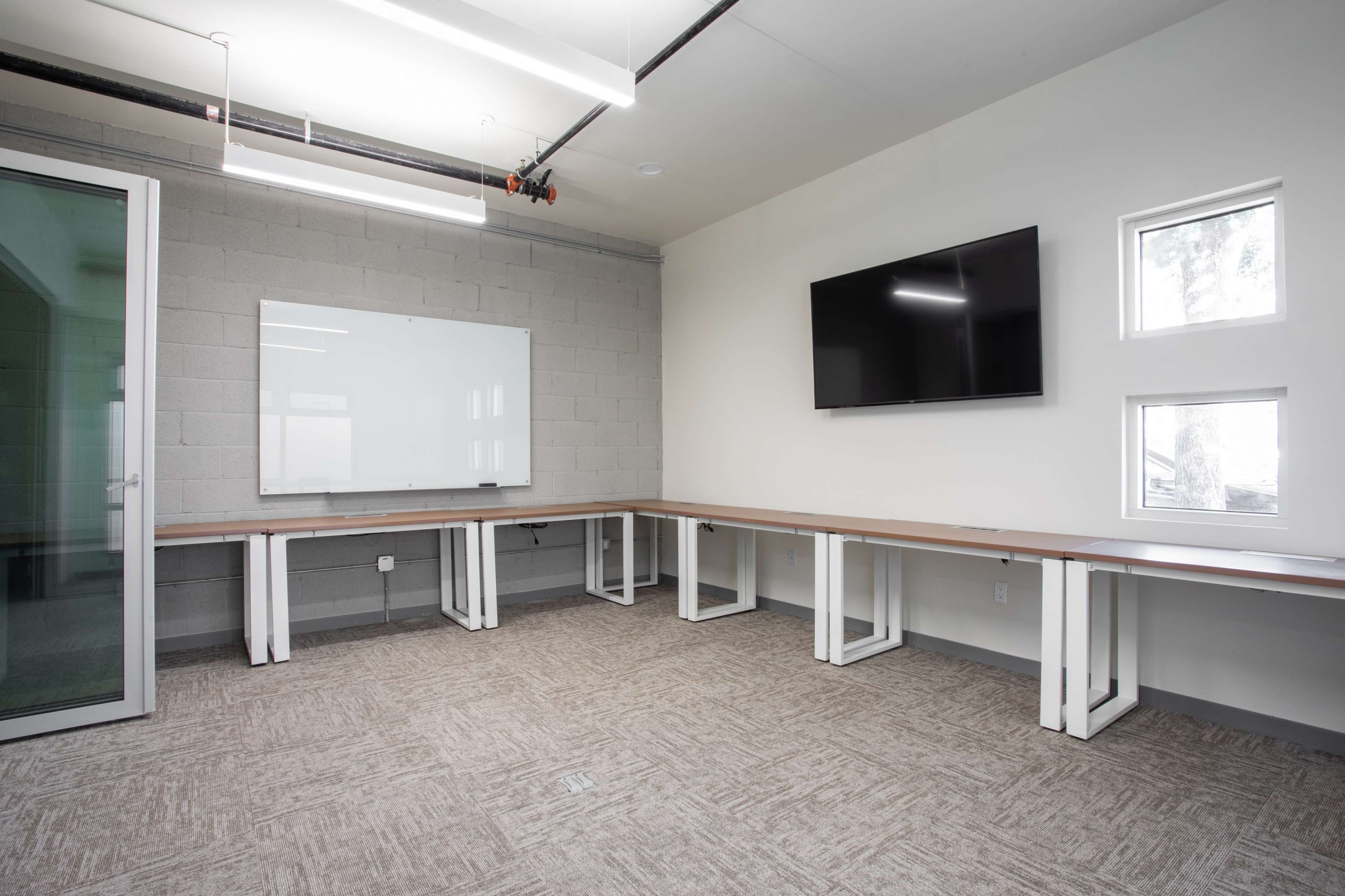 A modern conference room with a long table, a whiteboard, a television, and two windows, all within a minimalist design.