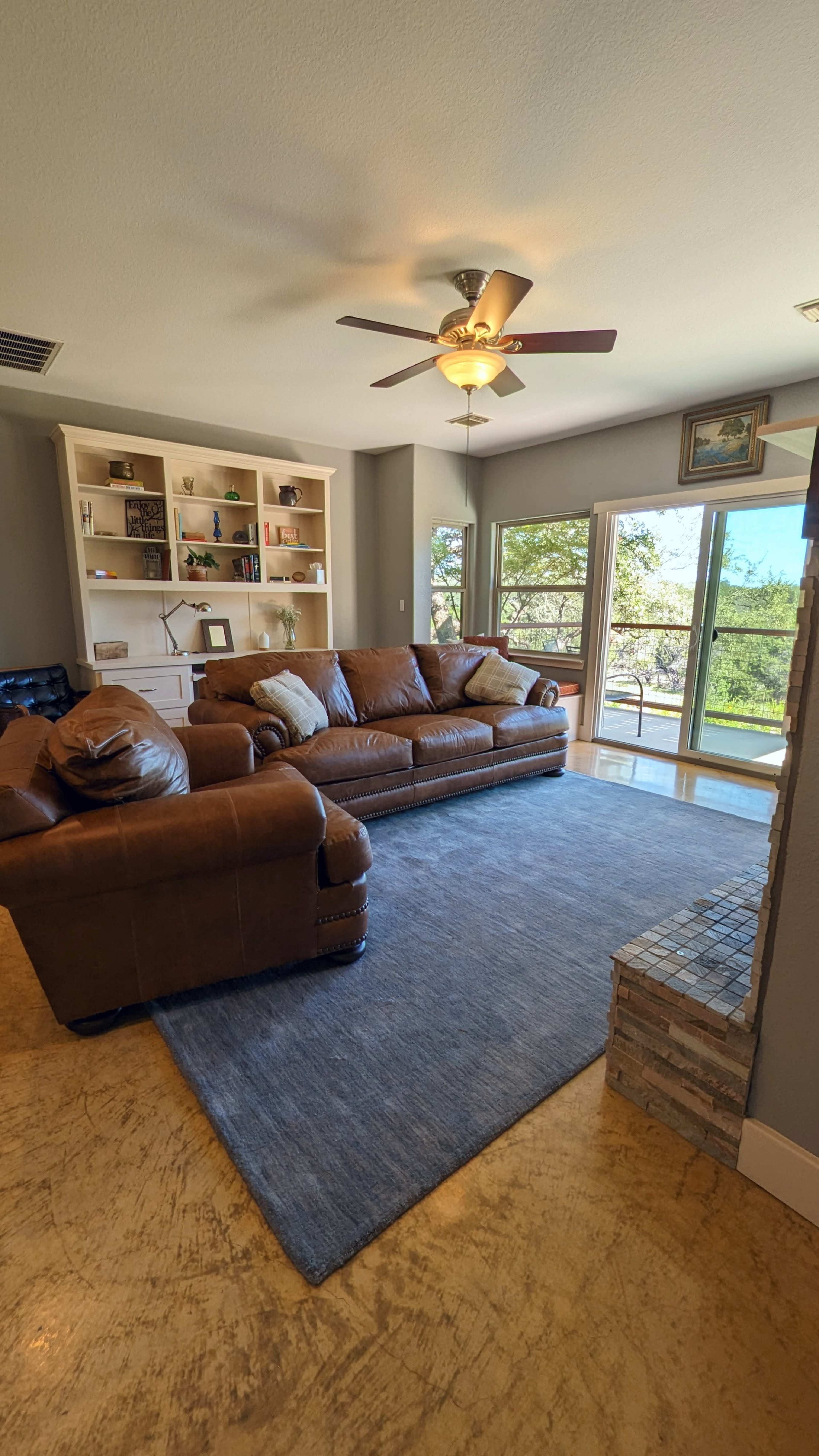 A living room featuring a brown leather sectional sofa, a light gray rug, a stone fireplace, and a large window with views of the outdoors.