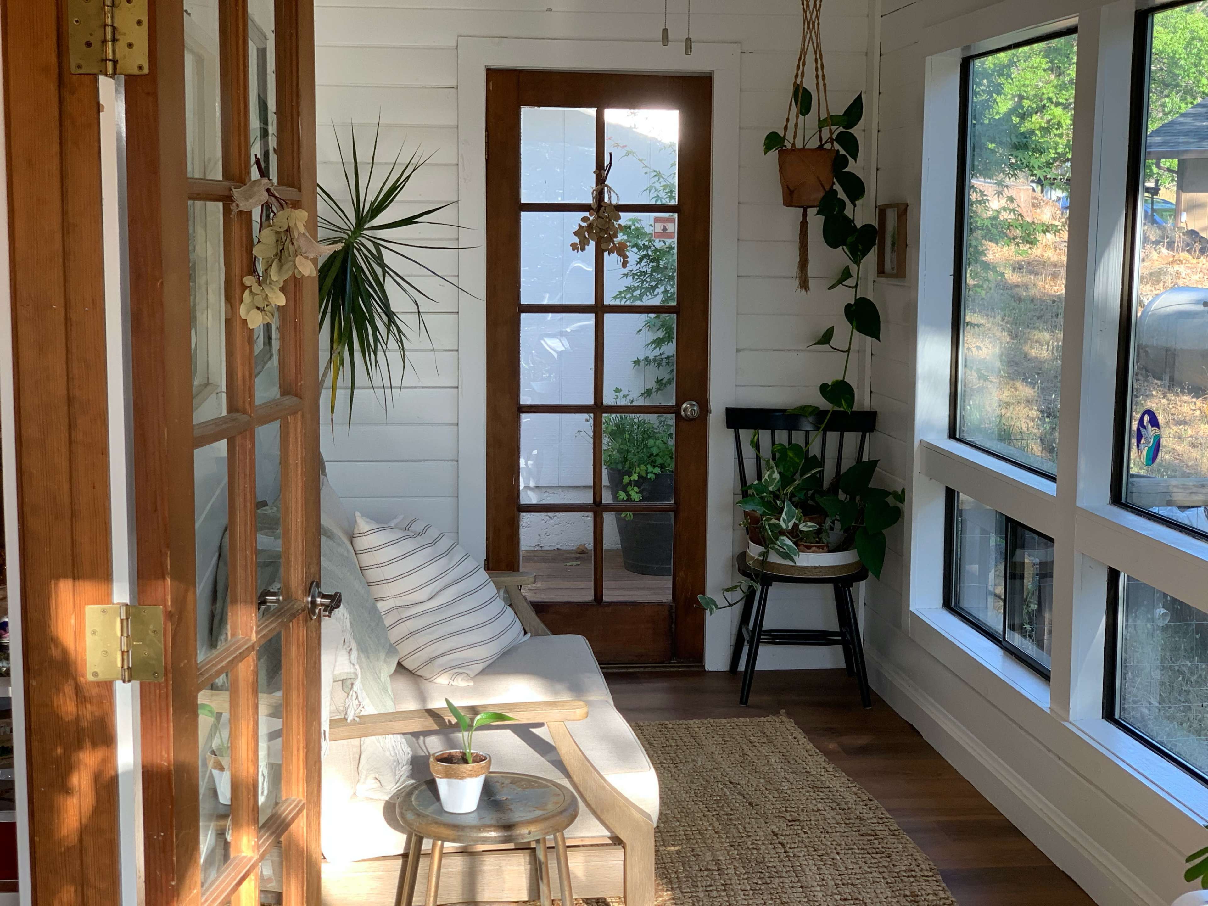 A cozy sunlit porch features a white couch, plants, and a glass door leading outside.