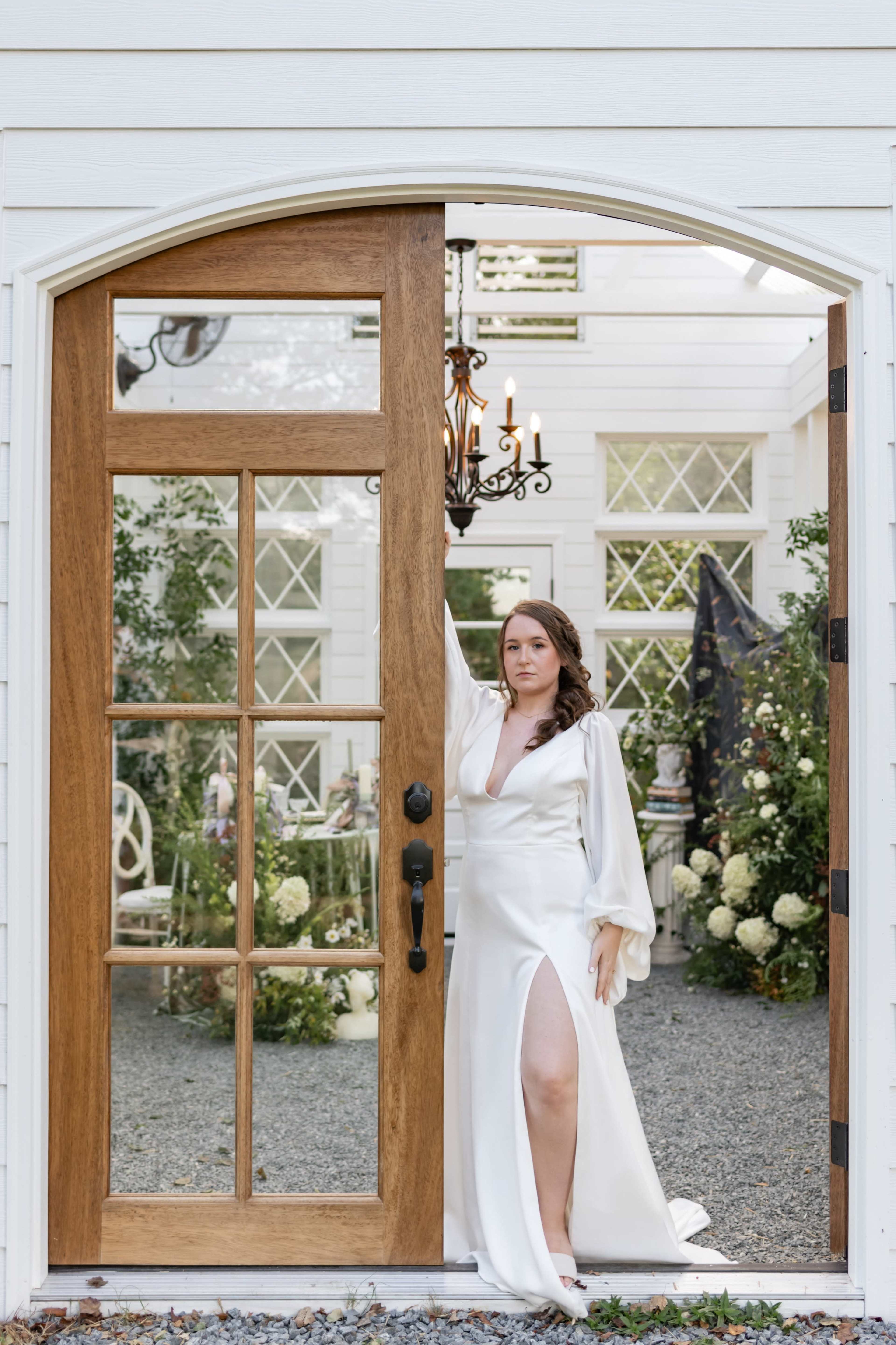 A woman in a white dress stands at an arched wooden door, holding onto the doorframe while looking into a garden setting adorned with greenery and flowers.
