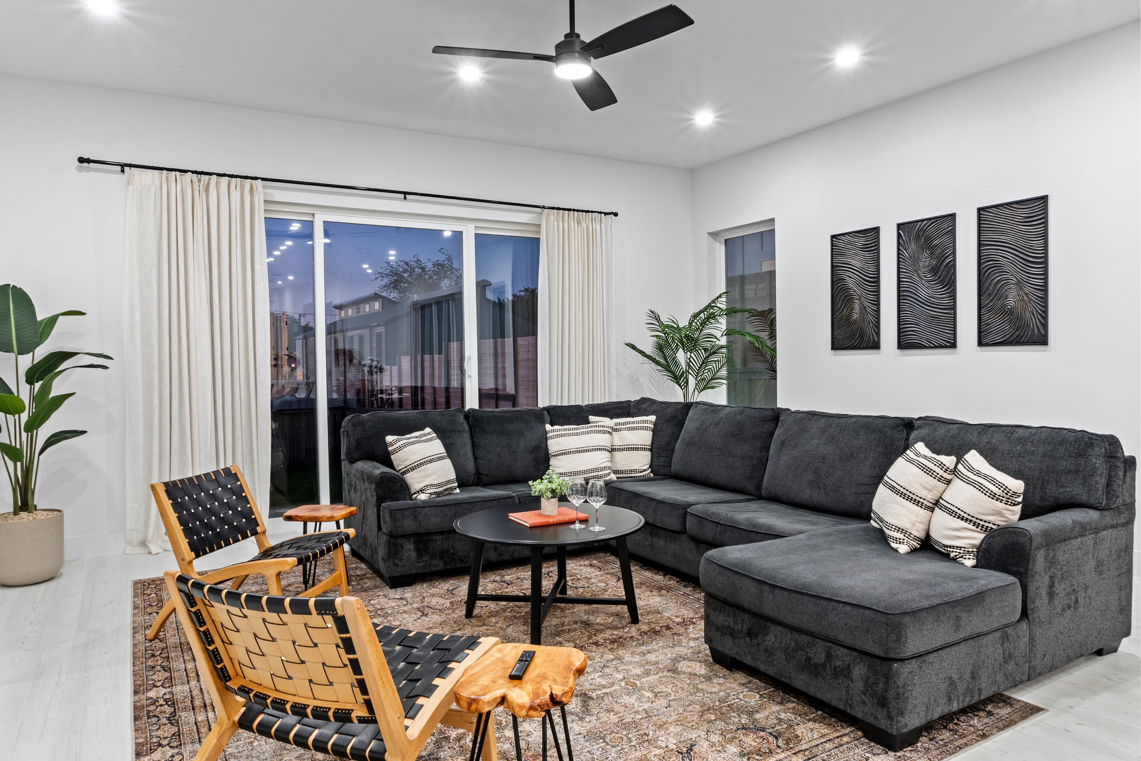 The image shows a modern living room featuring a dark sectional sofa, two wooden chairs, a round coffee table, and decorative wall art, all illuminated by ceiling lights.