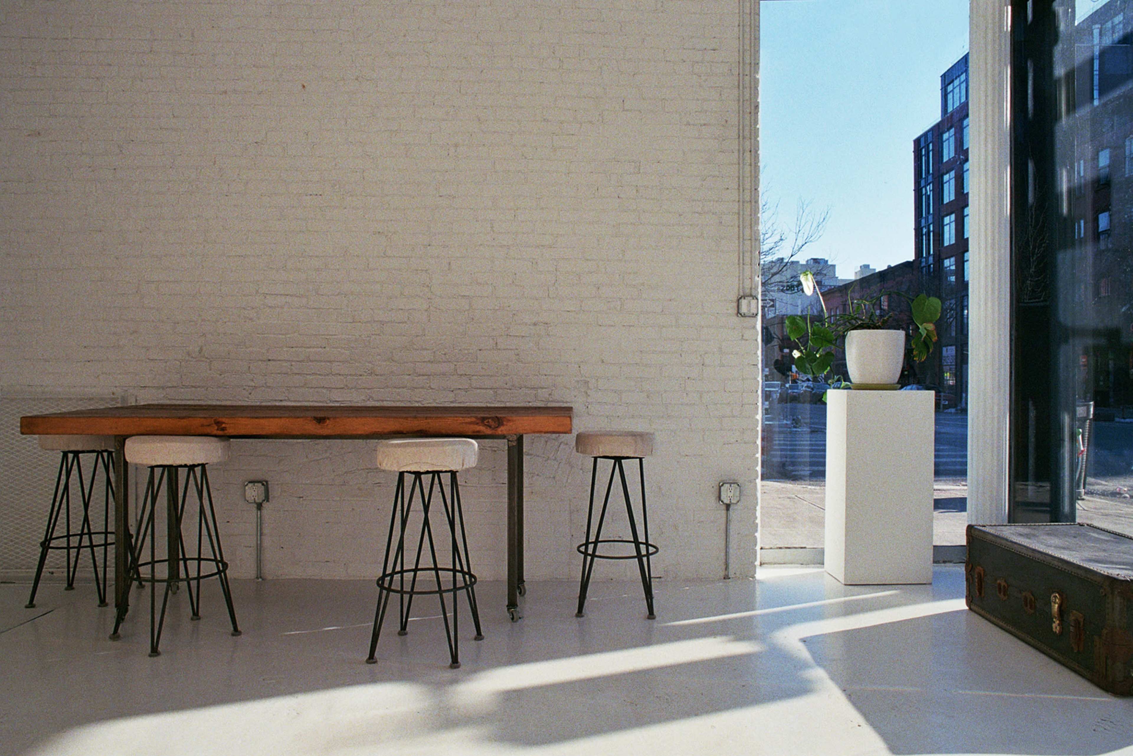 The image shows a modern, minimalistic interior featuring a wooden table with three metal stools, a white pedestal with a plant, and large windows letting in natural light.