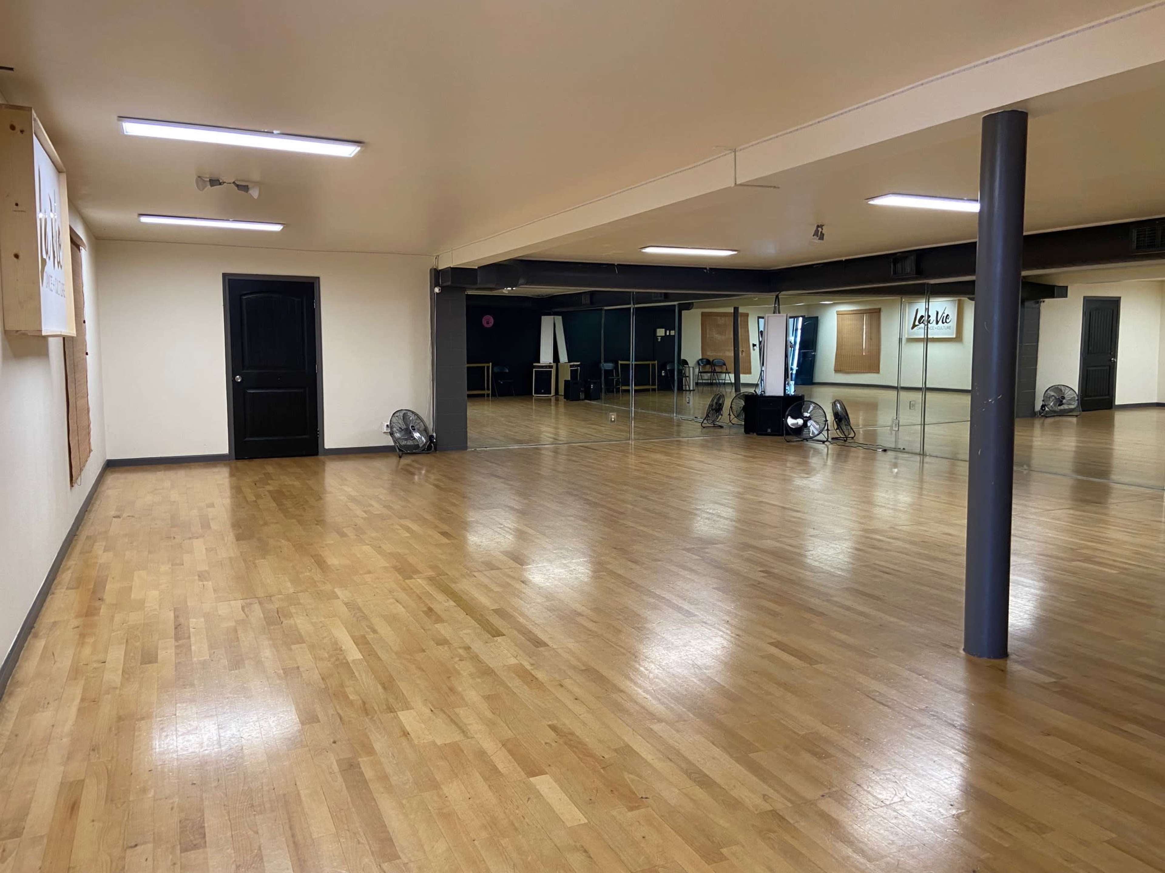 An empty dance studio with wooden floors, large mirrors on one wall, and a single black door.