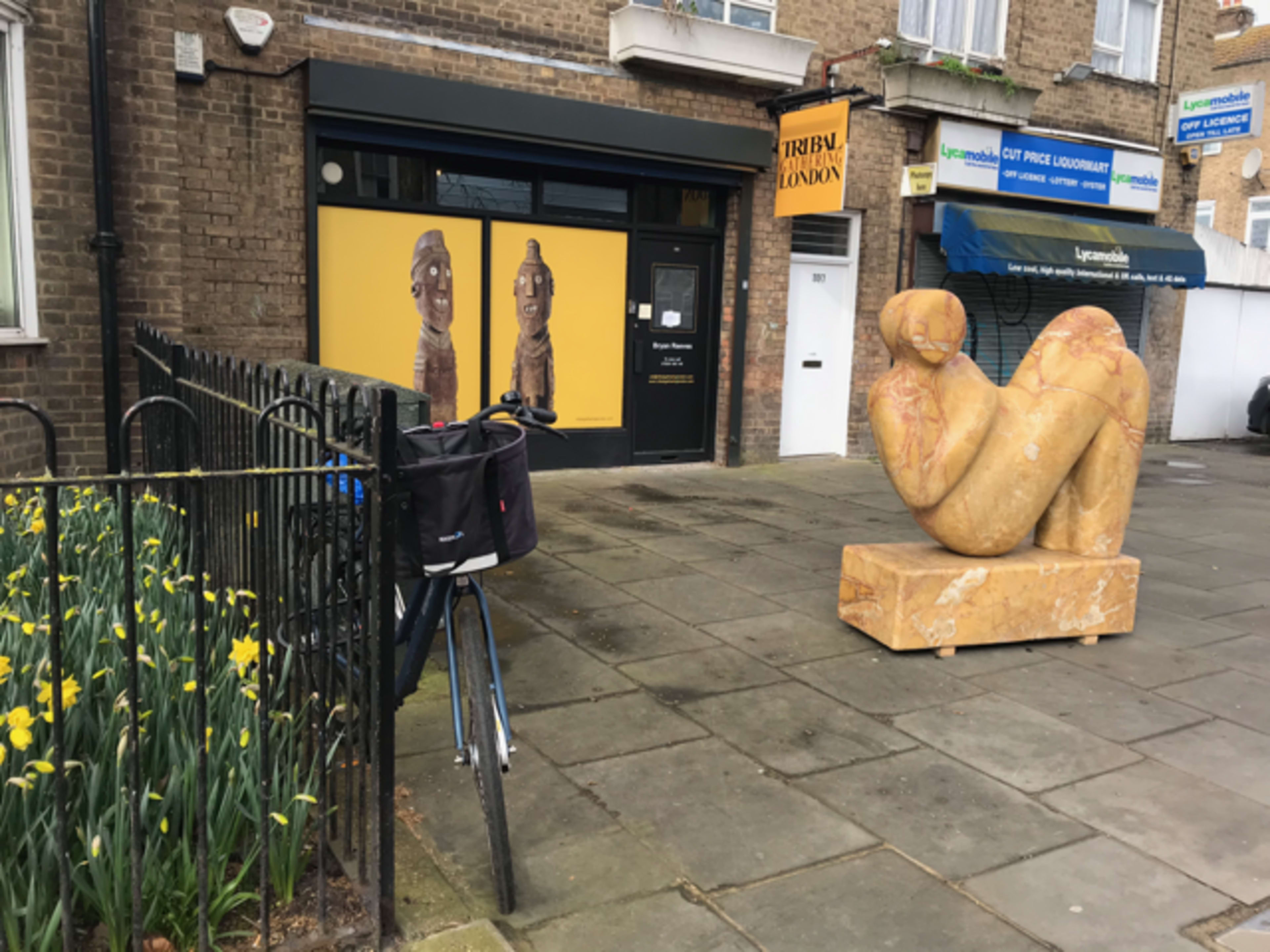The scene shows a bicycle parked next to a flowerbed with yellow daffodils, a stone sculpture of a figure on the sidewalk, and a shop front displaying tribal art.