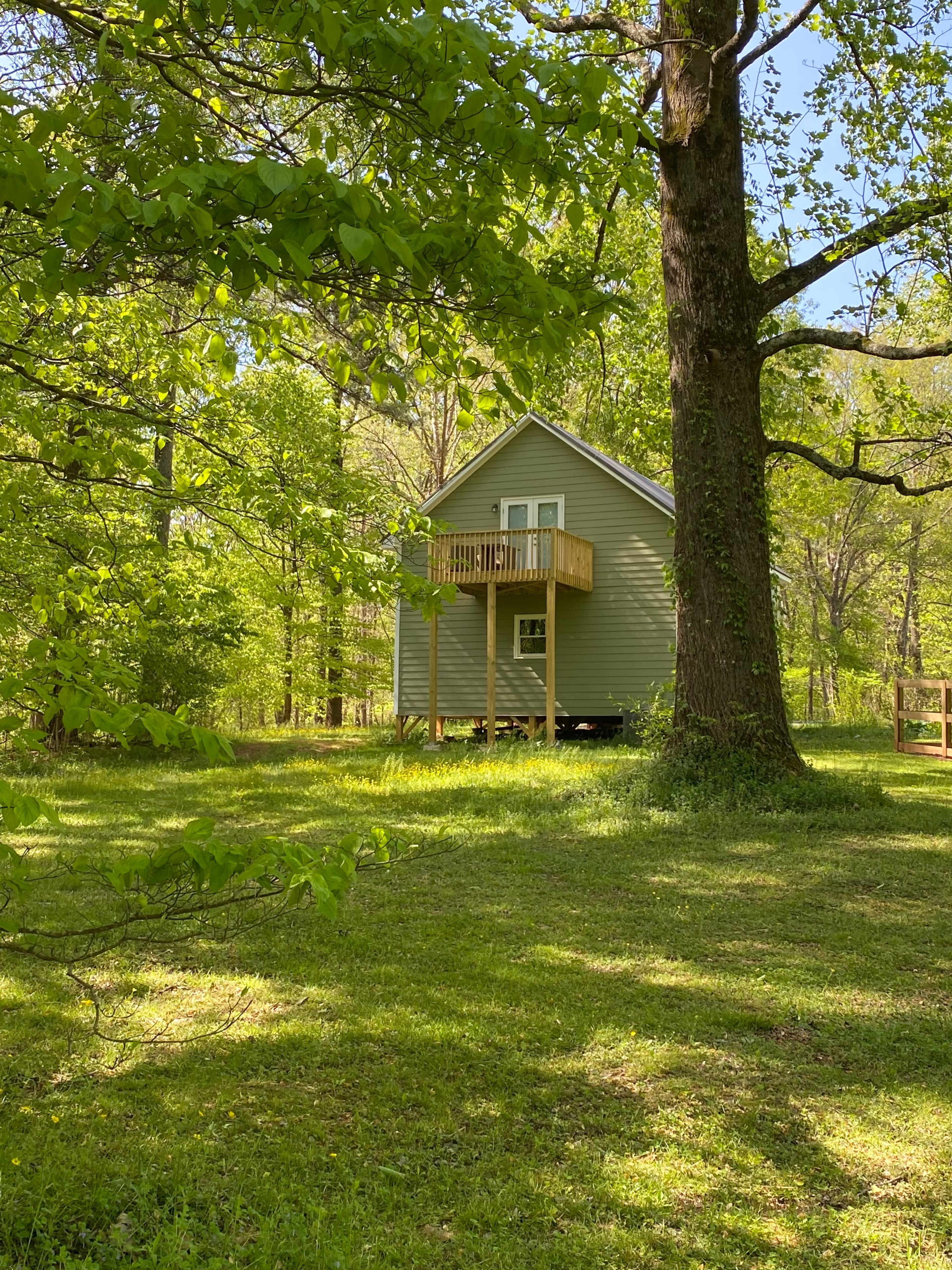 A small gray house with a balcony stands among lush green trees in a wooded area.