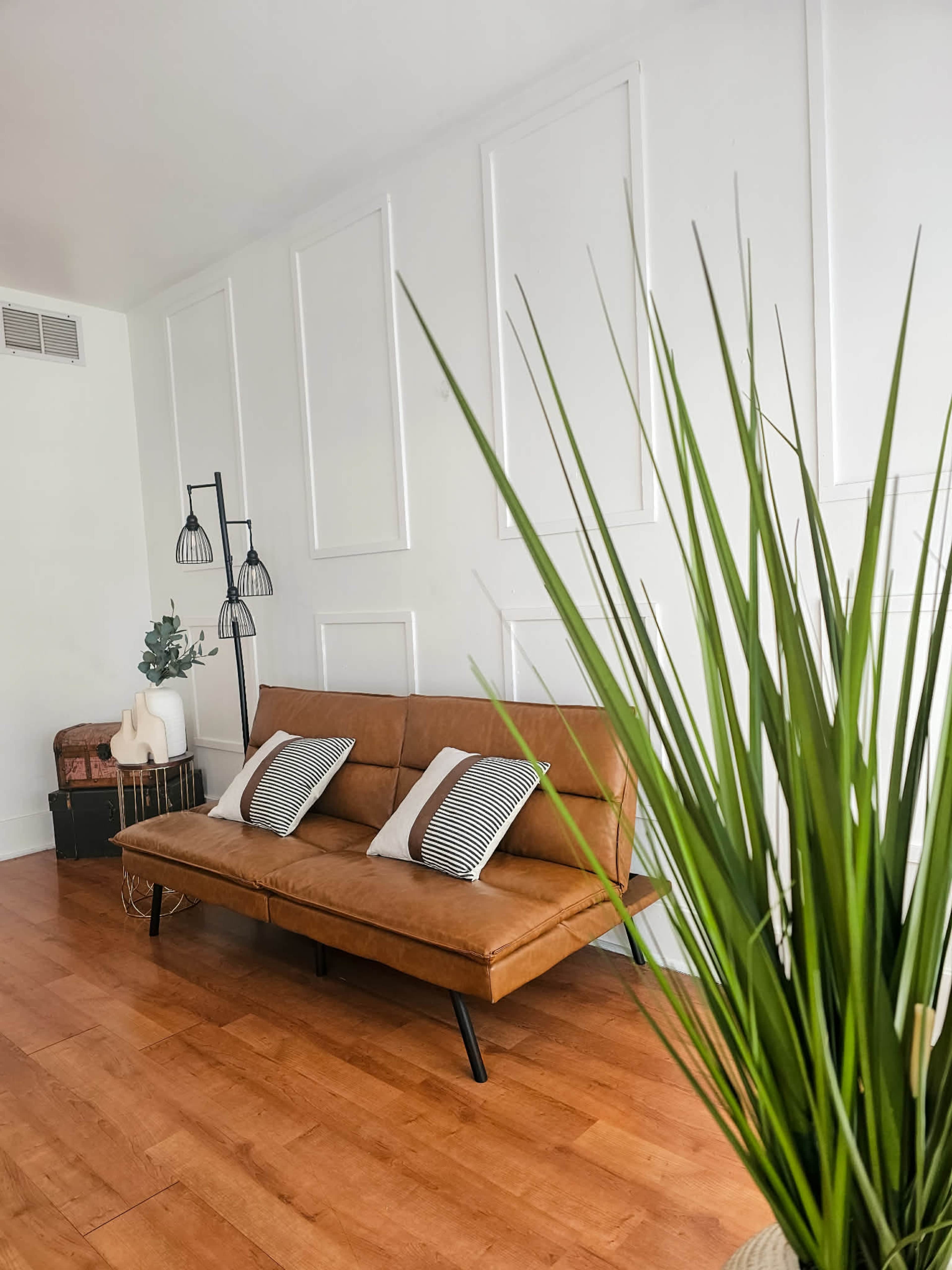 The image shows a modern living room with a brown sofa and decorative pillows, a tall plant in the foreground, and minimalist wall paneling behind.