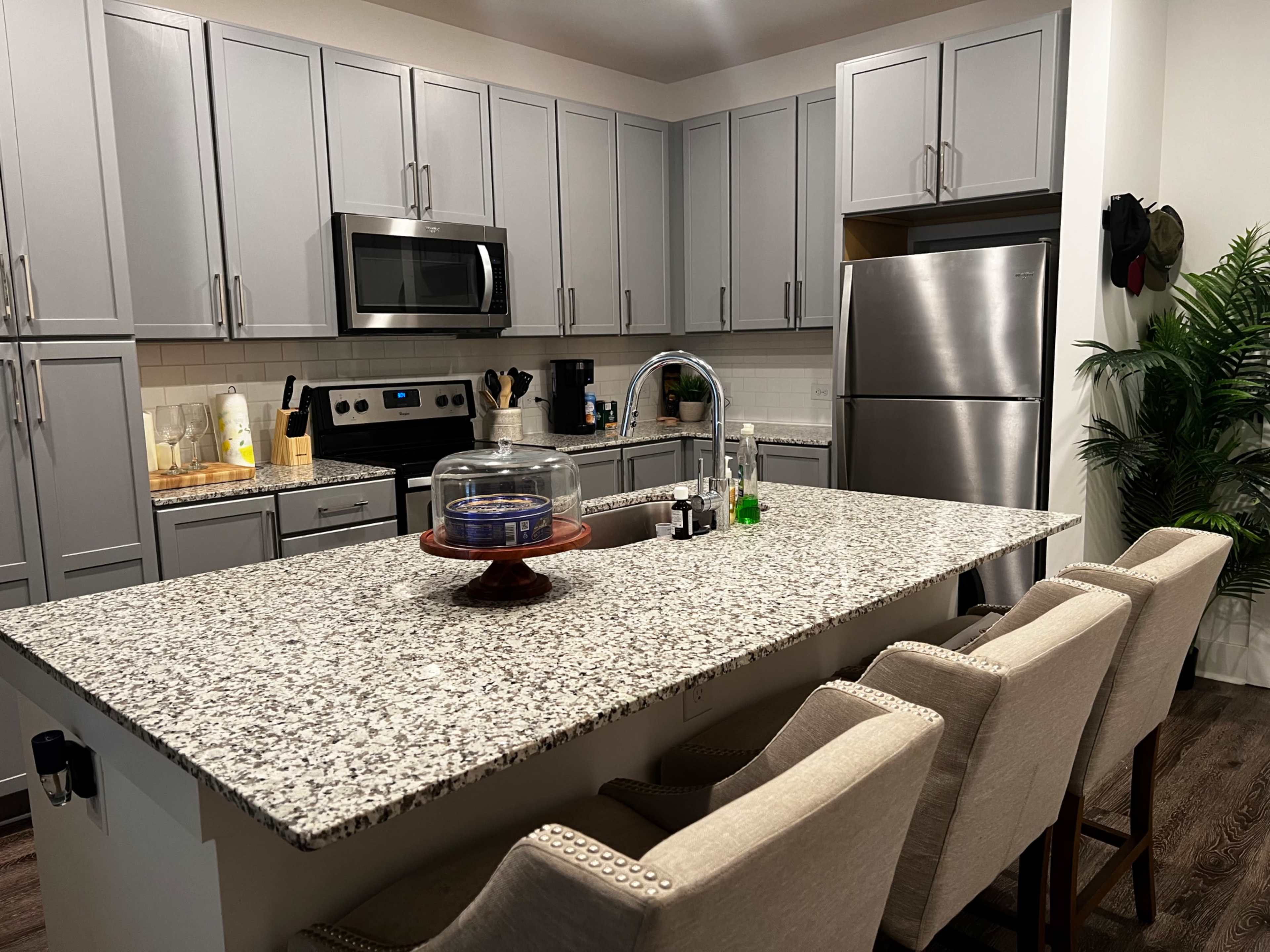 A modern kitchen with gray cabinets, a granite island, stainless steel appliances, and four upholstered bar stools.